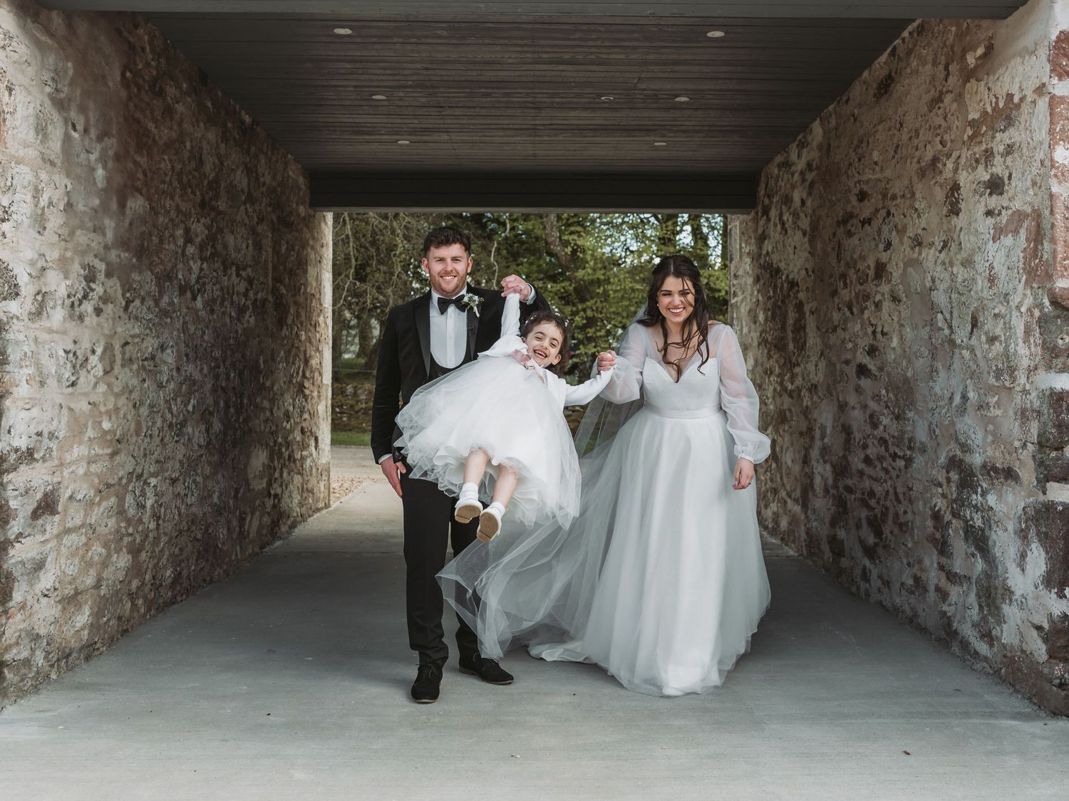Newlywed couple walks joyfully through a rustic stone tunnel in elegant wedding attire at Kinclune Steading.