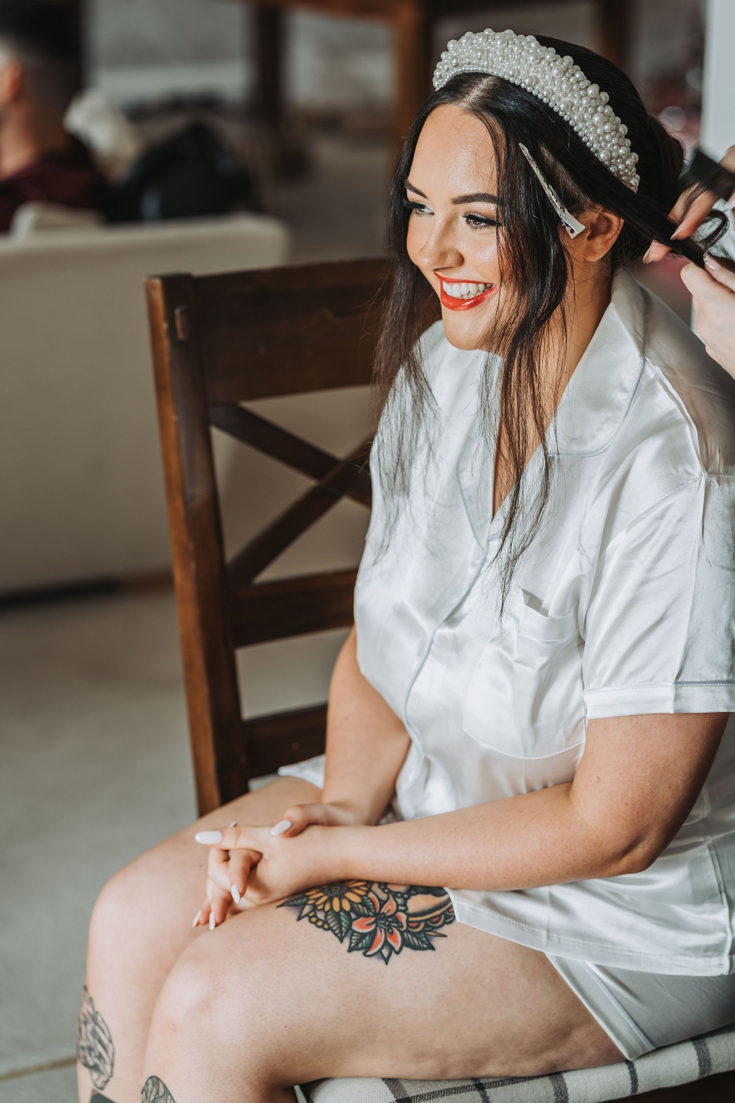 Bride in white robe and crystal headpiece smiles while getting ready, showing leg tattoos.