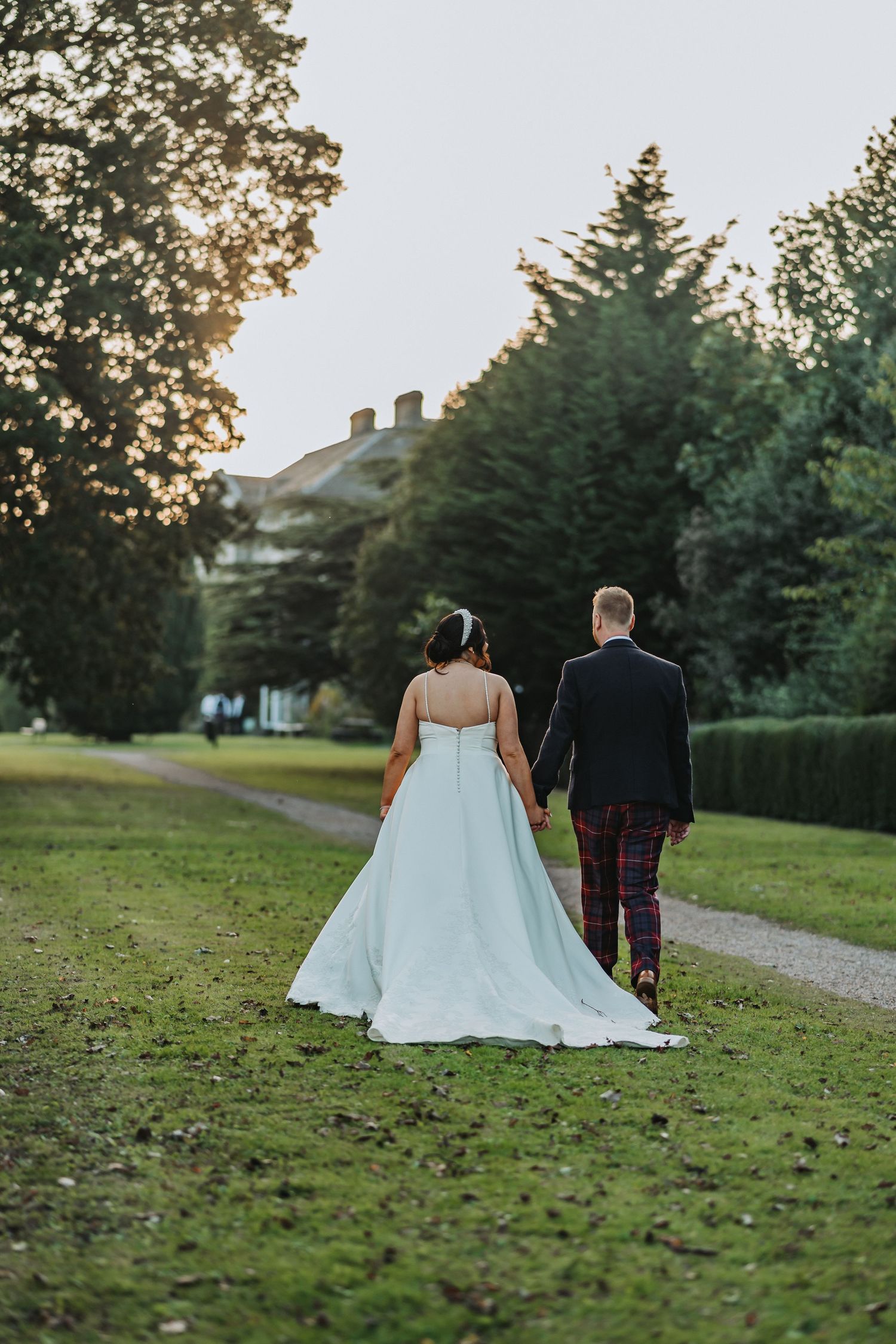 Bride and groom walk down garden path at sunset with elegant hedges and Oswald House in background.