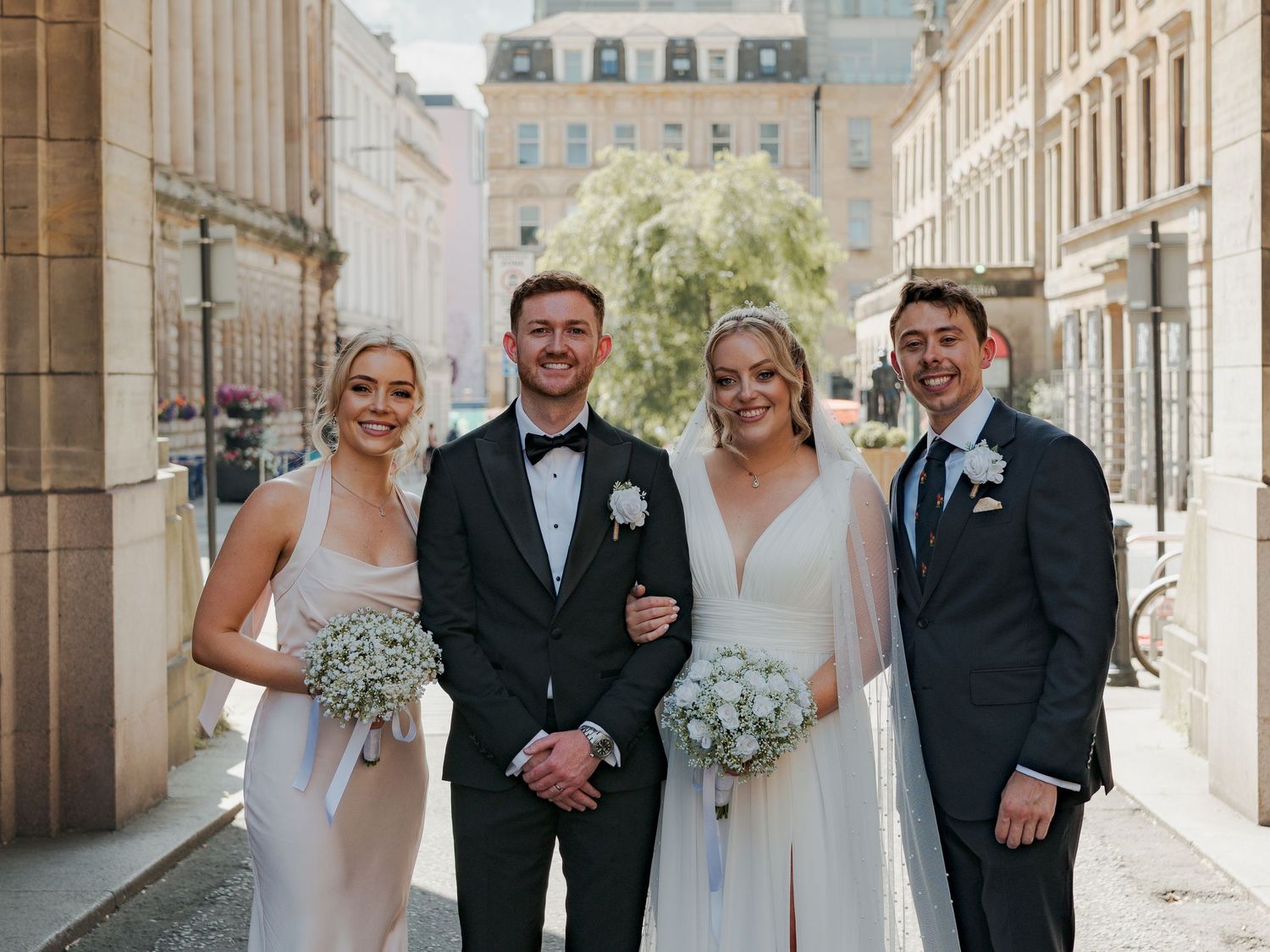 A wedding party poses together in formal attire along a stone building exterior with elegant Glasgow architecture.