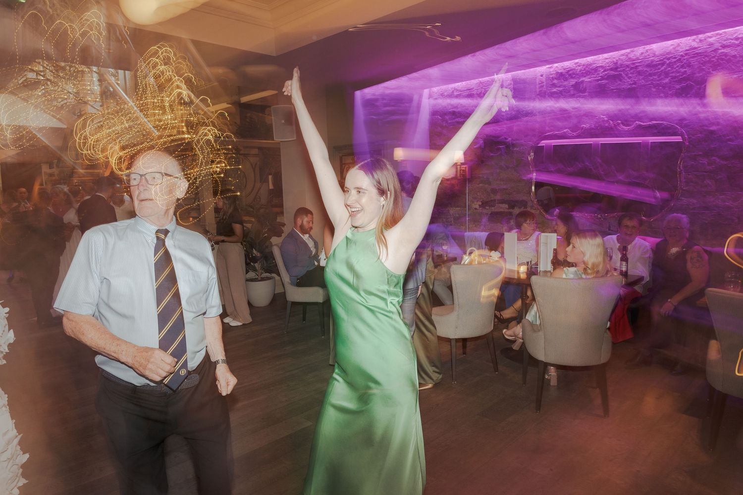 Wedding guests dancing under purple mood lighting at an indoor reception venue.