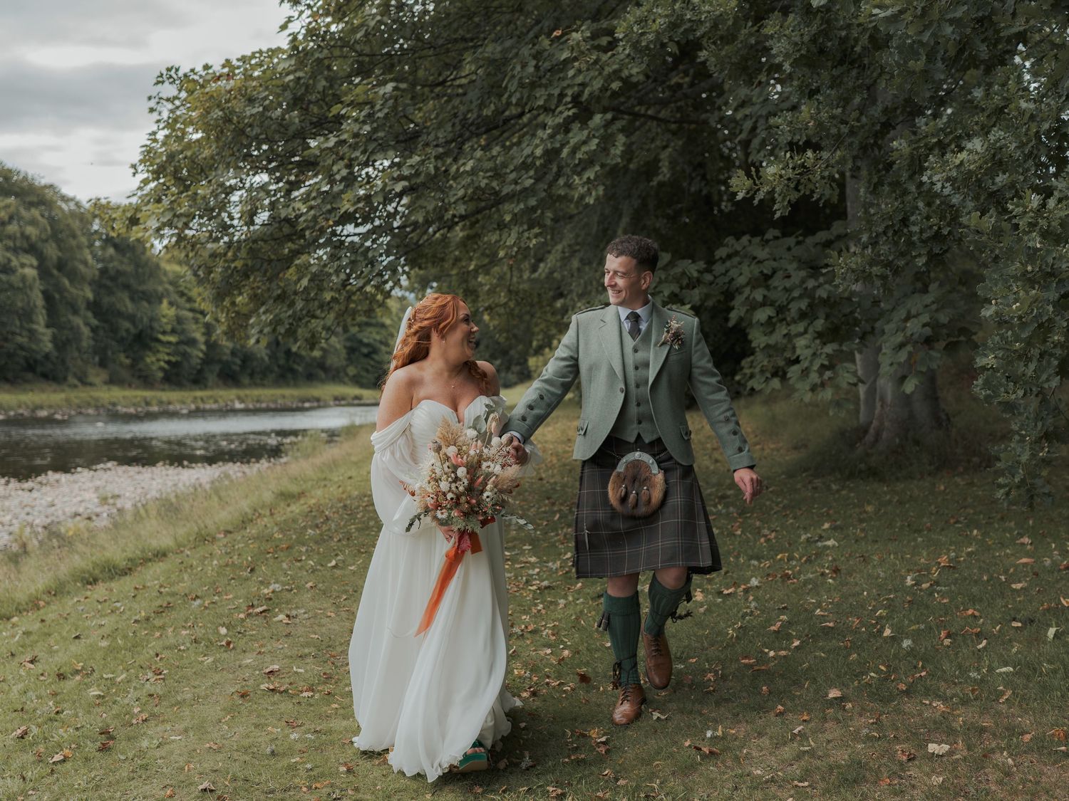 A couple in wedding attire walk hand in hand along a river bank at Banchory Lodge, with the groom wearing a traditional kilt.