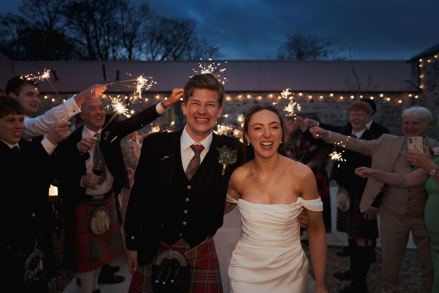 Bride and groom walk through sparkler send-off at twilight, groom in Scottish tartan kilt, at romantic outdoor wedding celebration.