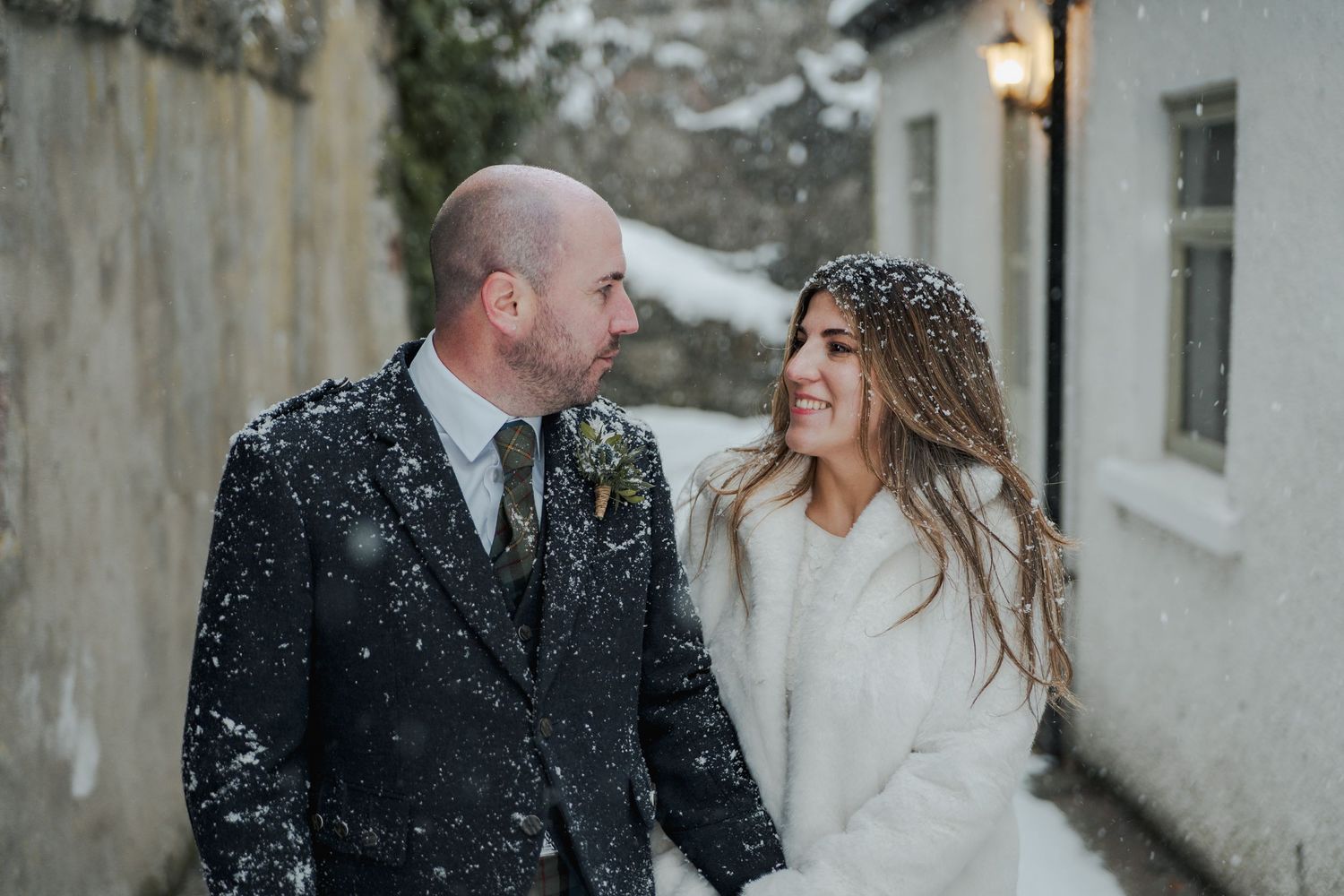 Bride and groom smiling at each other in falling snow during a romantic winter wedding outdoors.
