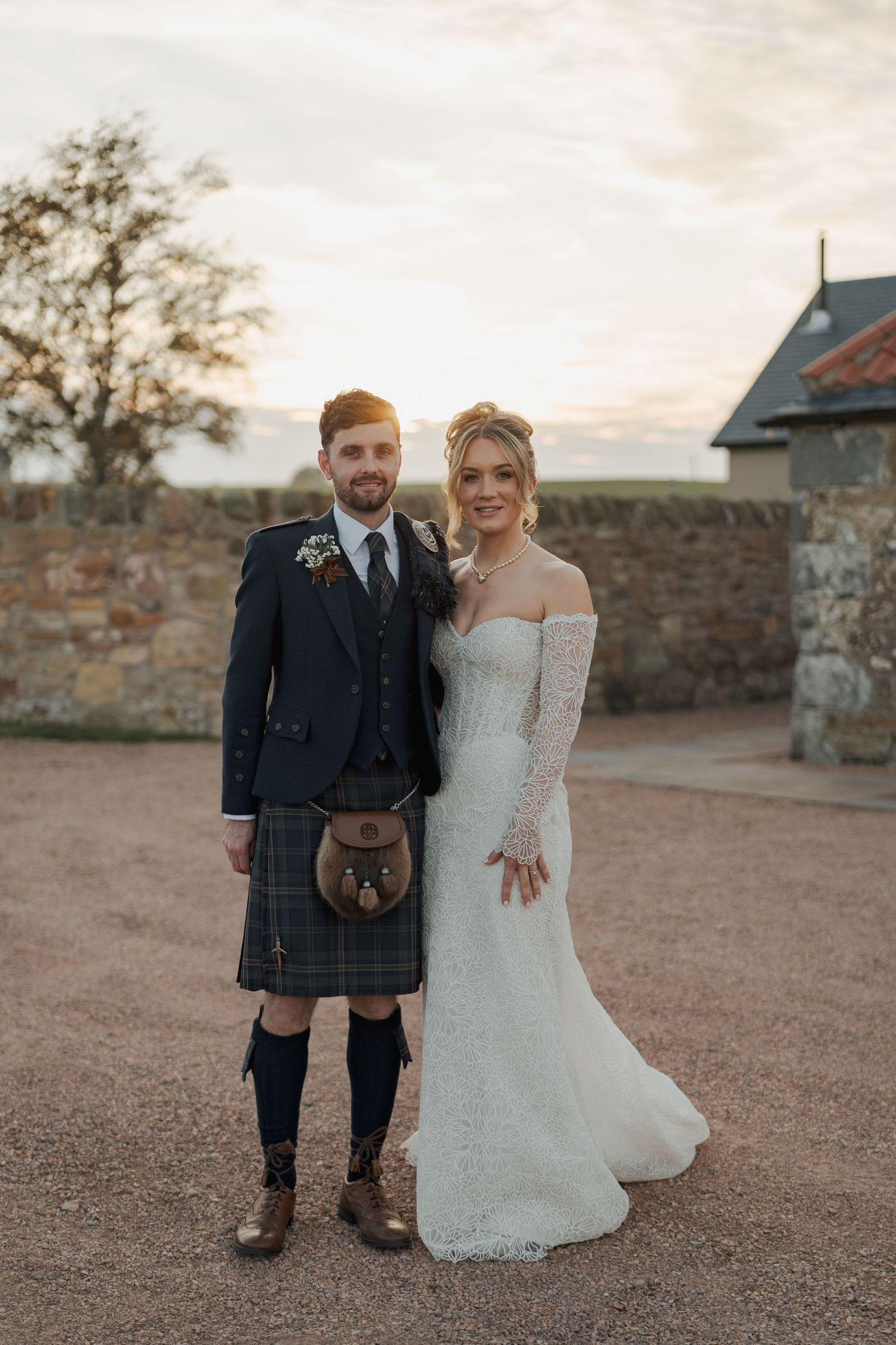 Couple in traditional Scottish wedding attire pose together at sunset against stone wall backdrop at Falside Mill.