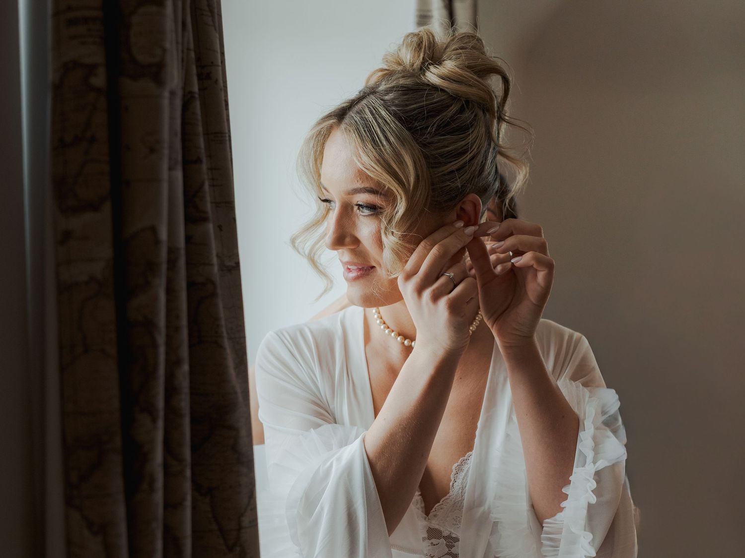 A bride in a white robe adjusts her hair while getting ready in front of a window with natural light streaming in.