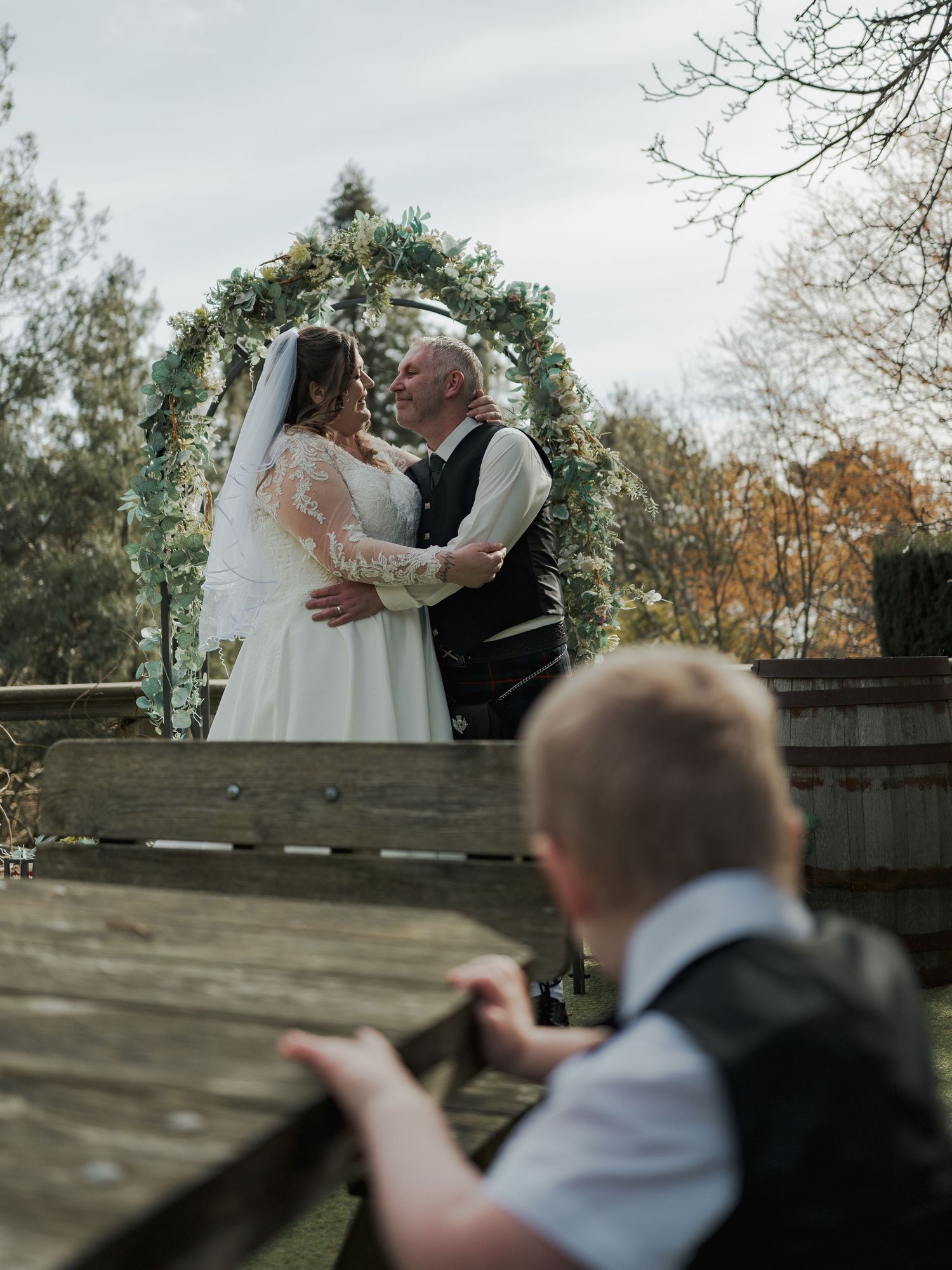 Bride and groom share a romantic kiss under a floral arch at an outdoor wedding ceremony.