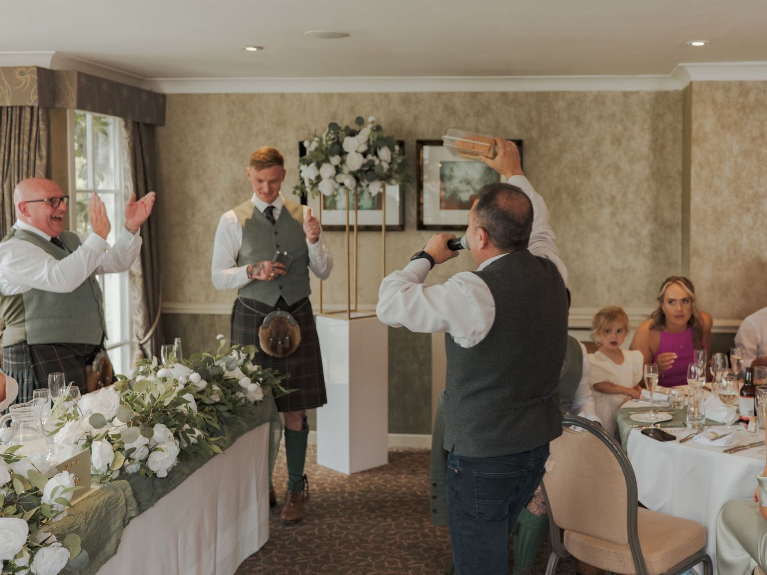 People gather in an elegant beige reception hall decorated with white floral arrangements for a wedding celebration.