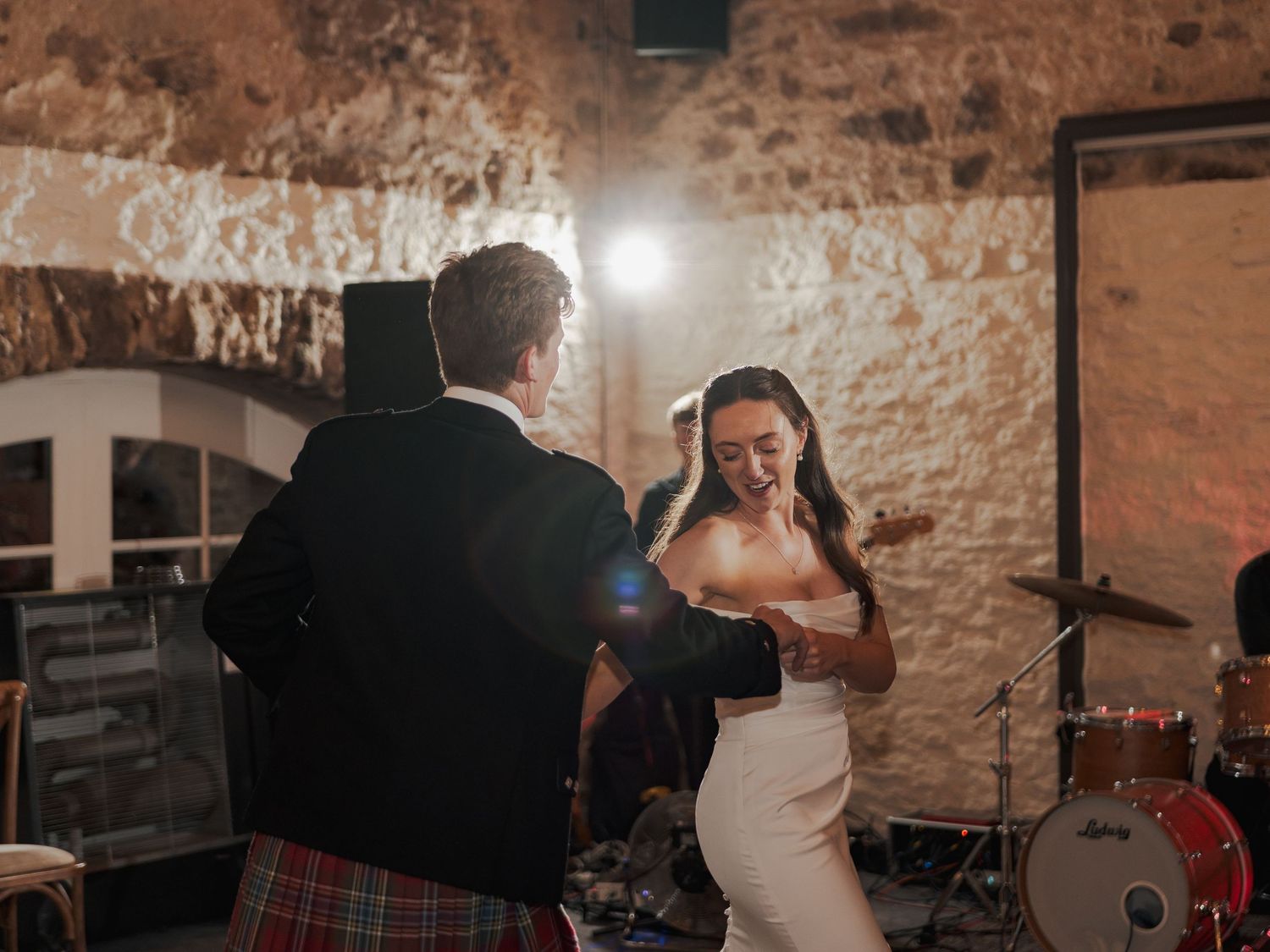 Bride and groom share first dance in rustic stone venue, groom wearing traditional Scottish tartan kilt.