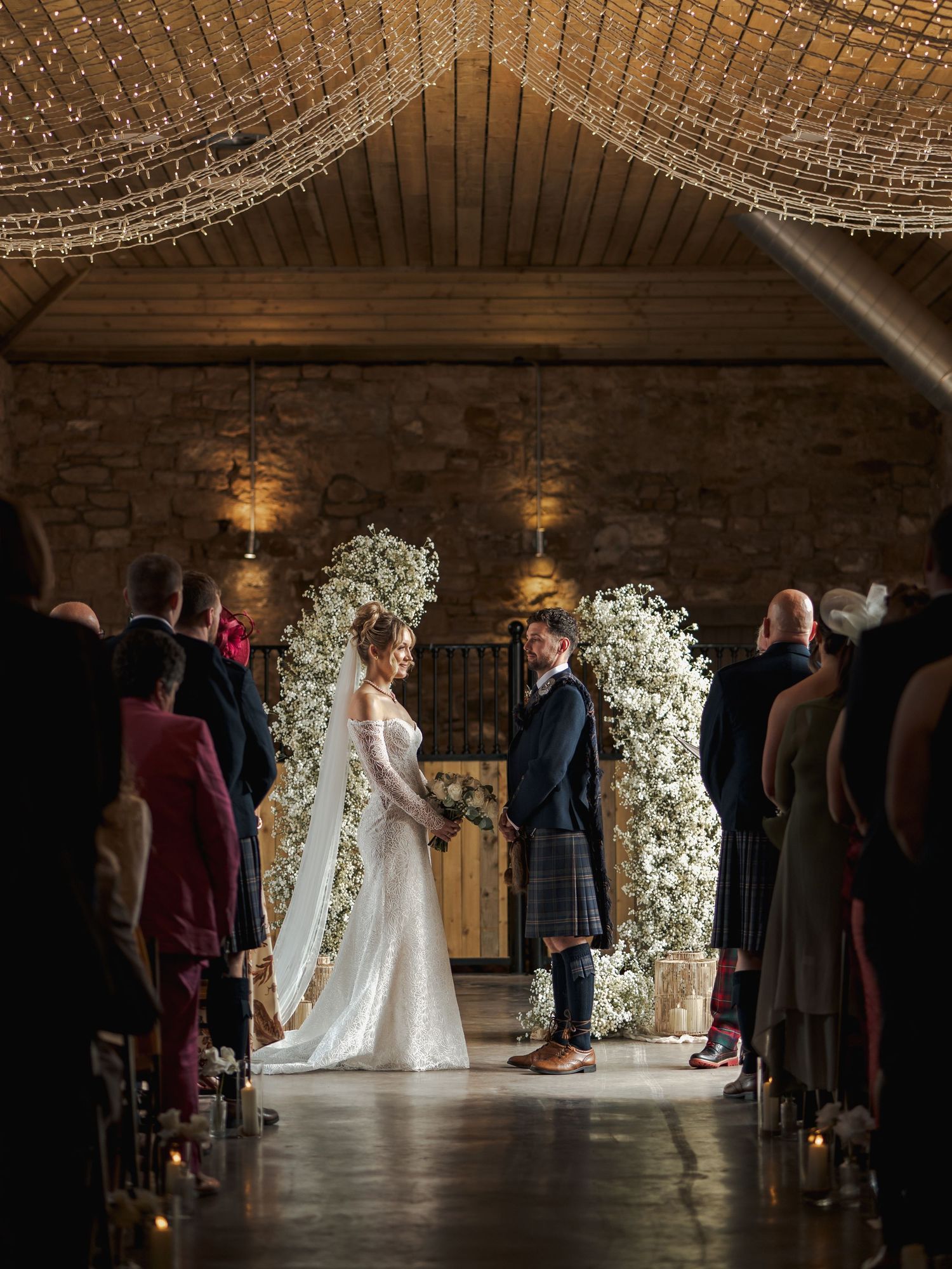 Wedding ceremony at Falside Mill with floral arch and guests standing in silhouette against natural light.