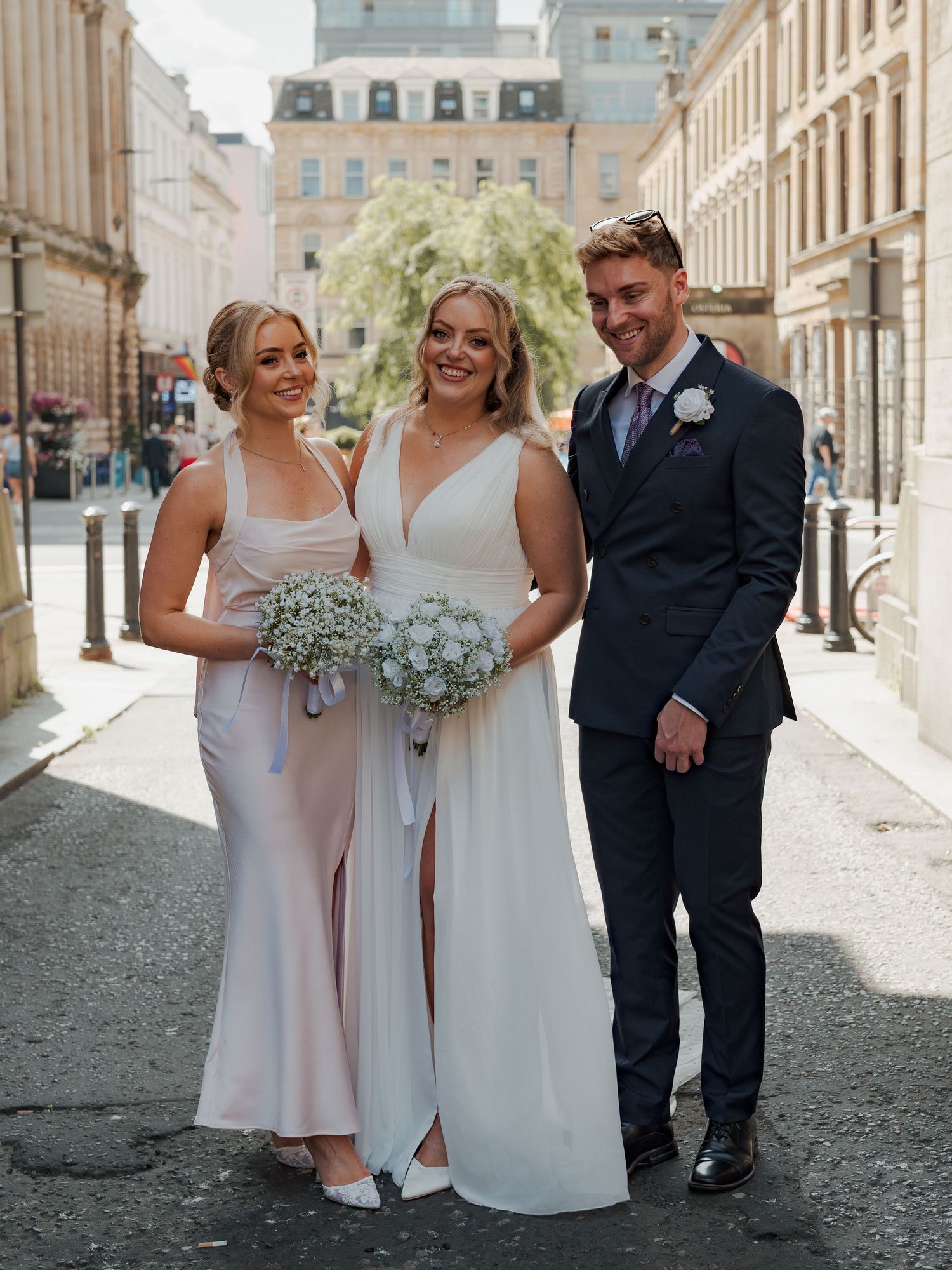 Wedding party members in pink and white dresses pose with groomsmen in suits on a charming Glasgow cobblestone street.