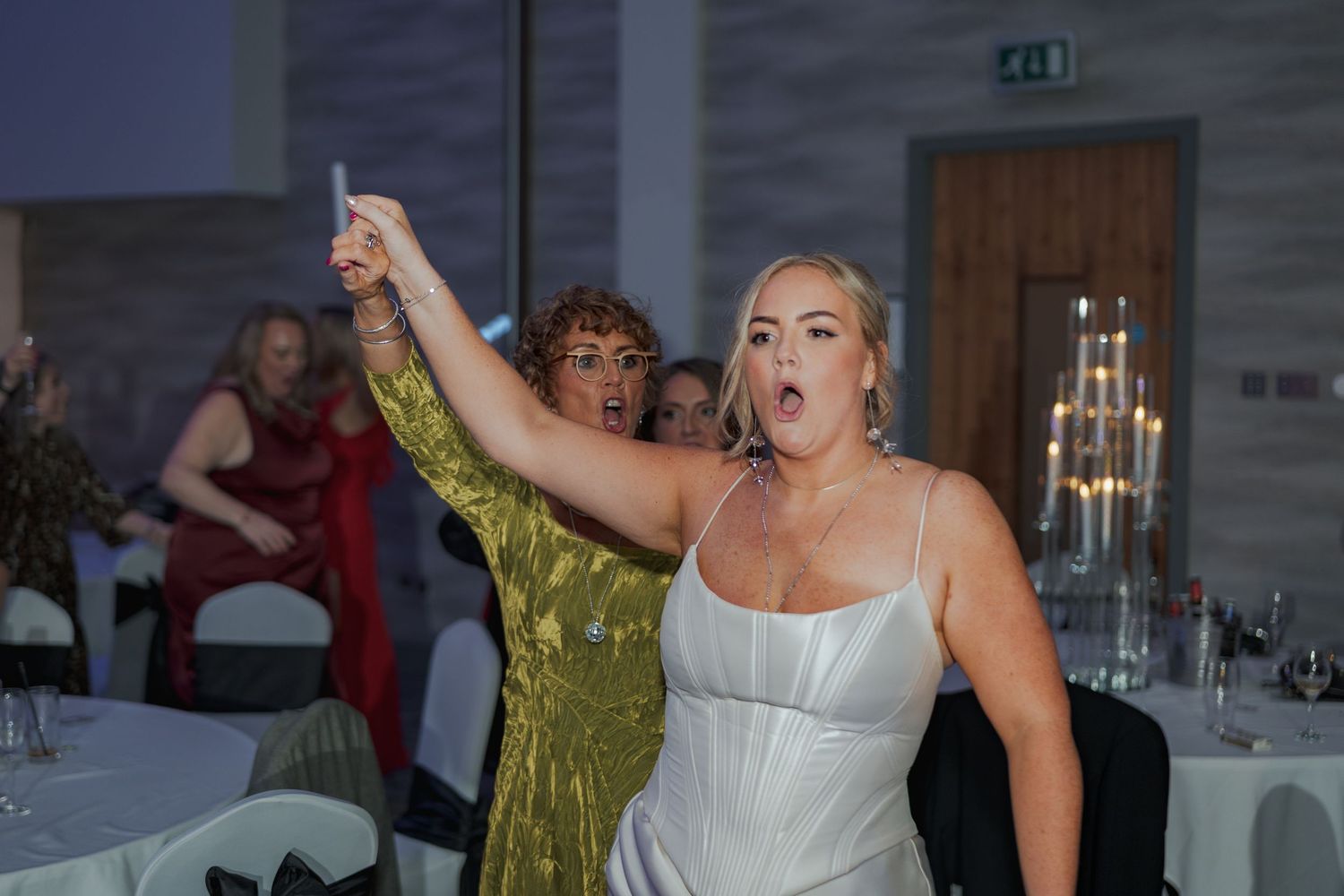 Wedding guests dancing and celebrating on a dance floor at an indoor reception venue with white decor and candlelight.