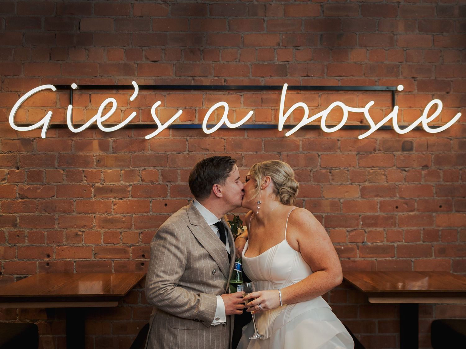 A couple shares a romantic kiss against a rustic brick wall backdrop at a wedding celebration.