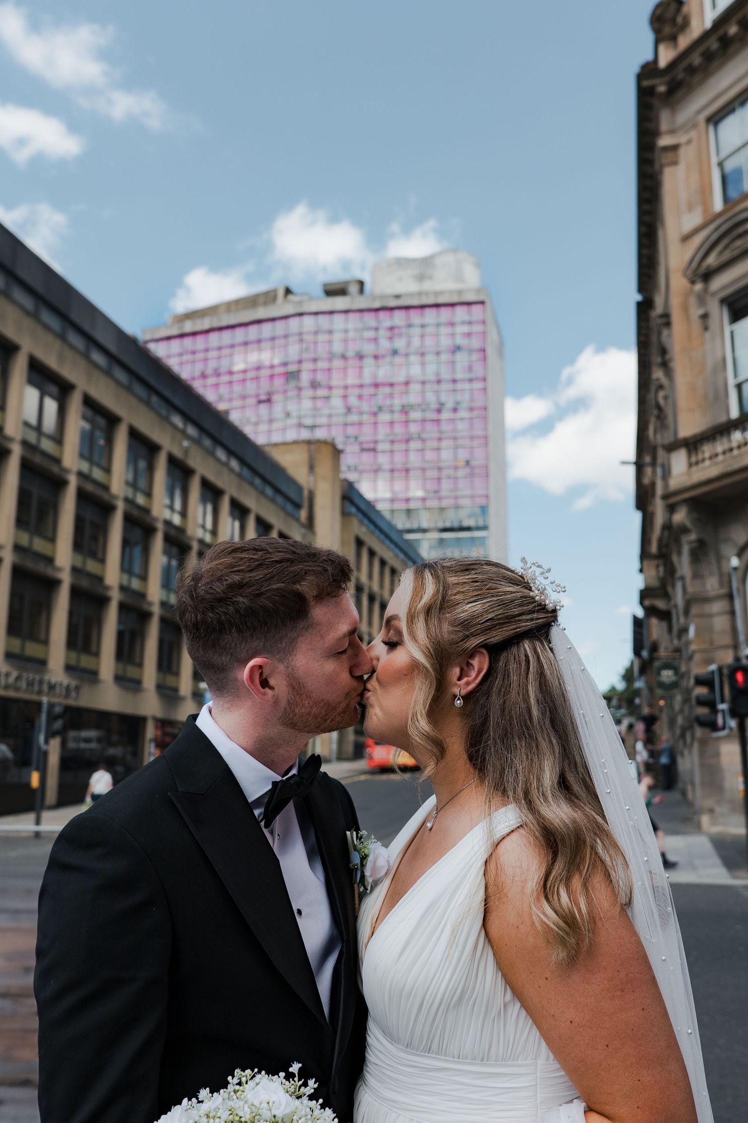 A newly married couple shares an intimate kiss on a city street with modern buildings and blue skies in the background.