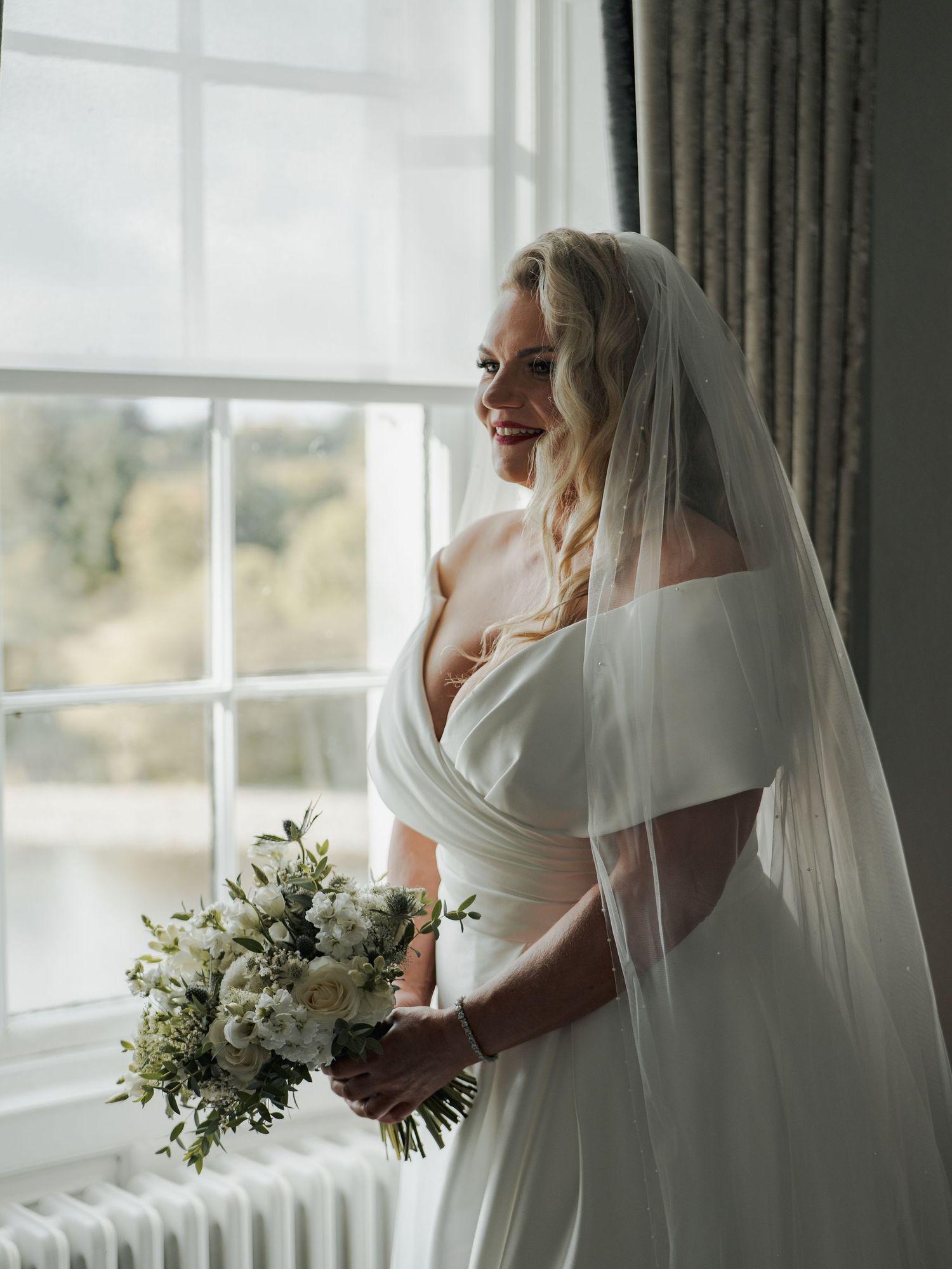 Bride in off-shoulder white gown and veil holds bouquet by window, smiling softly in elegant venue.