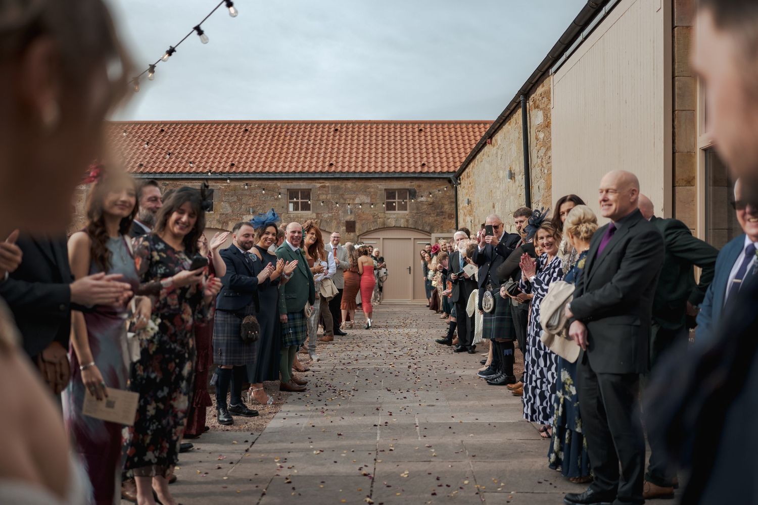 A wedding ceremony crowd stands in two rows creating an aisle outside Falside Mill.