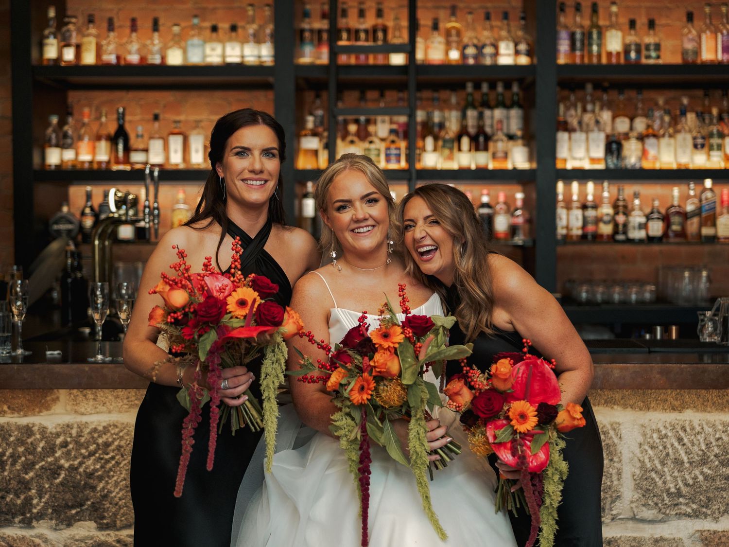 Wedding party members laugh and smile while holding vibrant red and orange bouquets at the Sandman Signature, Aberdeen.