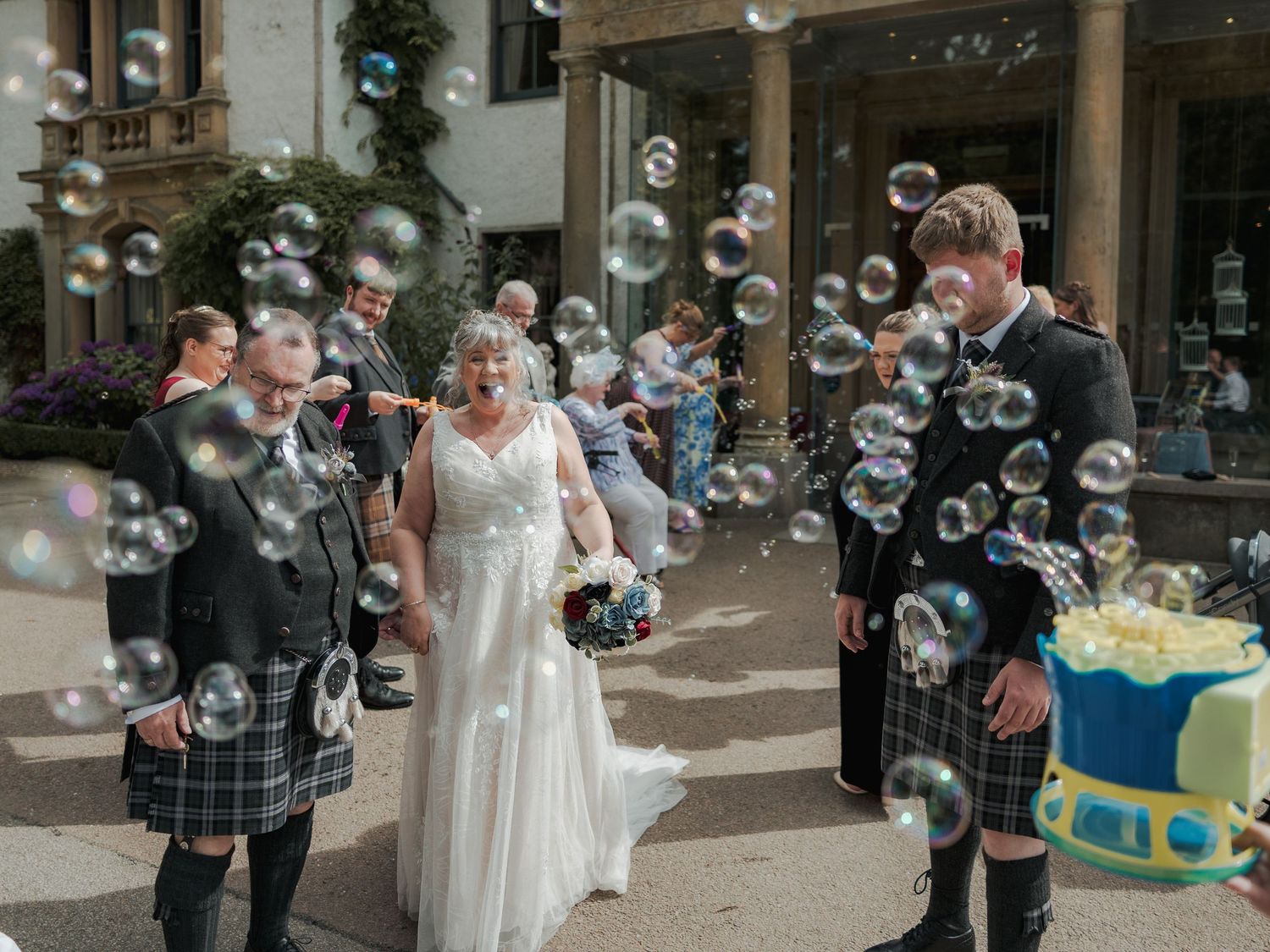 Wedding couple walks through bubbles outside venue, groom in tartan kilt, bride in white gown, guests celebrating behind them.