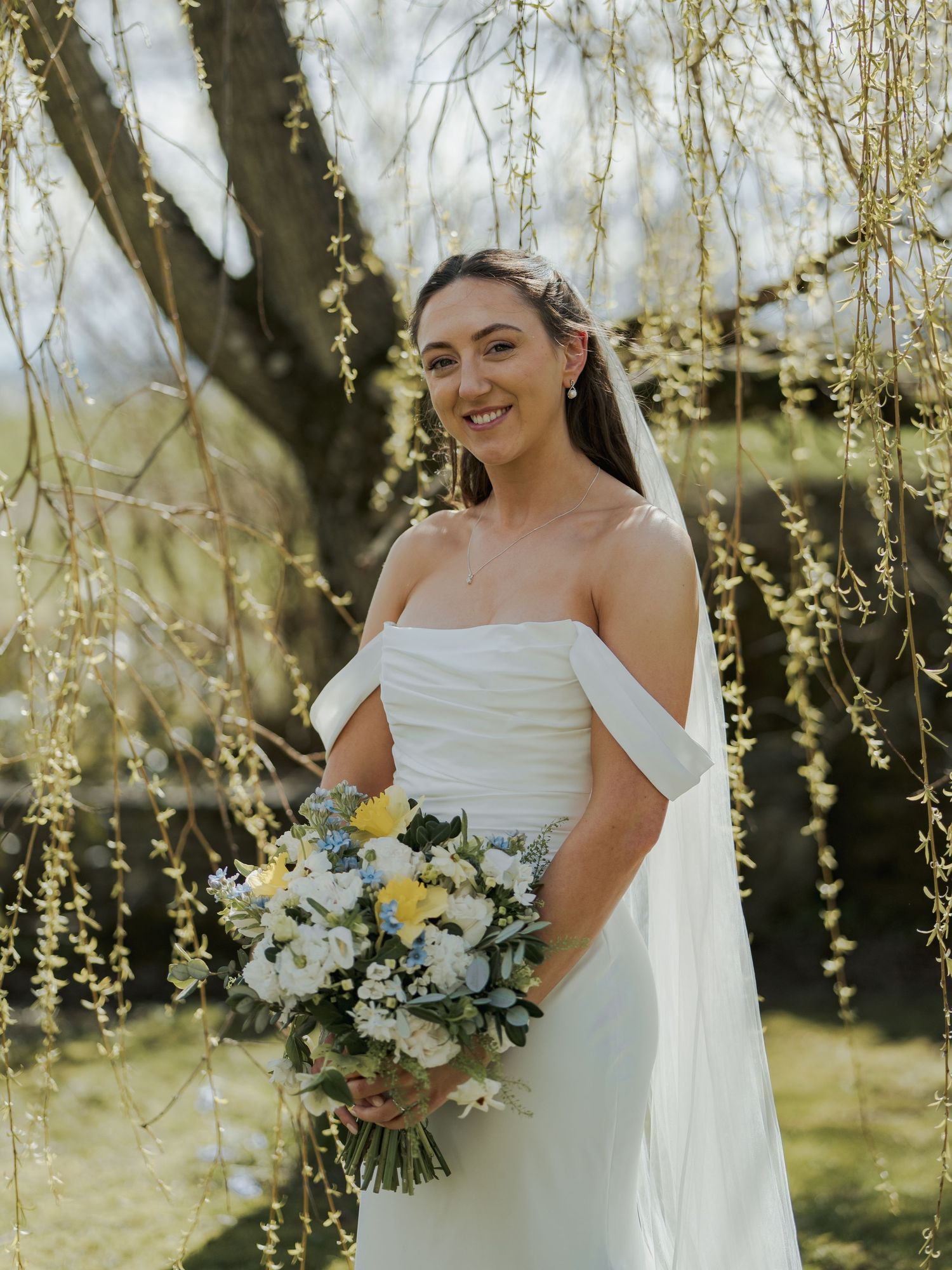 Smiling bride in off-shoulder white gown holds yellow and white bouquet under weeping willow tree branches.