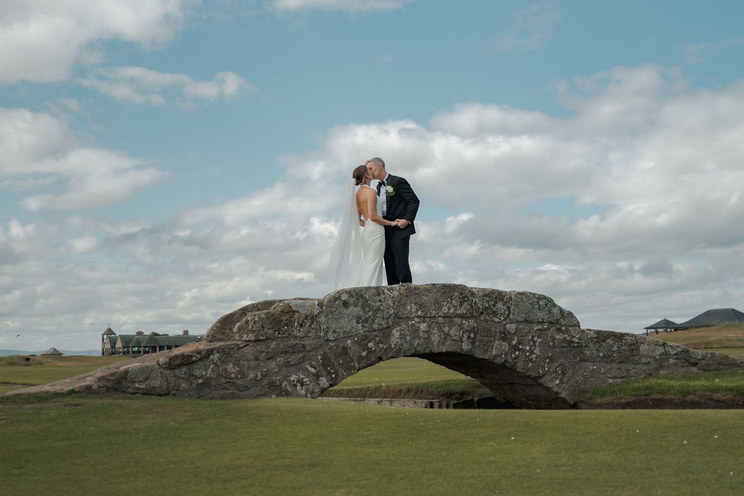 Bride and groom kissing on a stone bridge at a golf course under a cloudy sky on their wedding day.