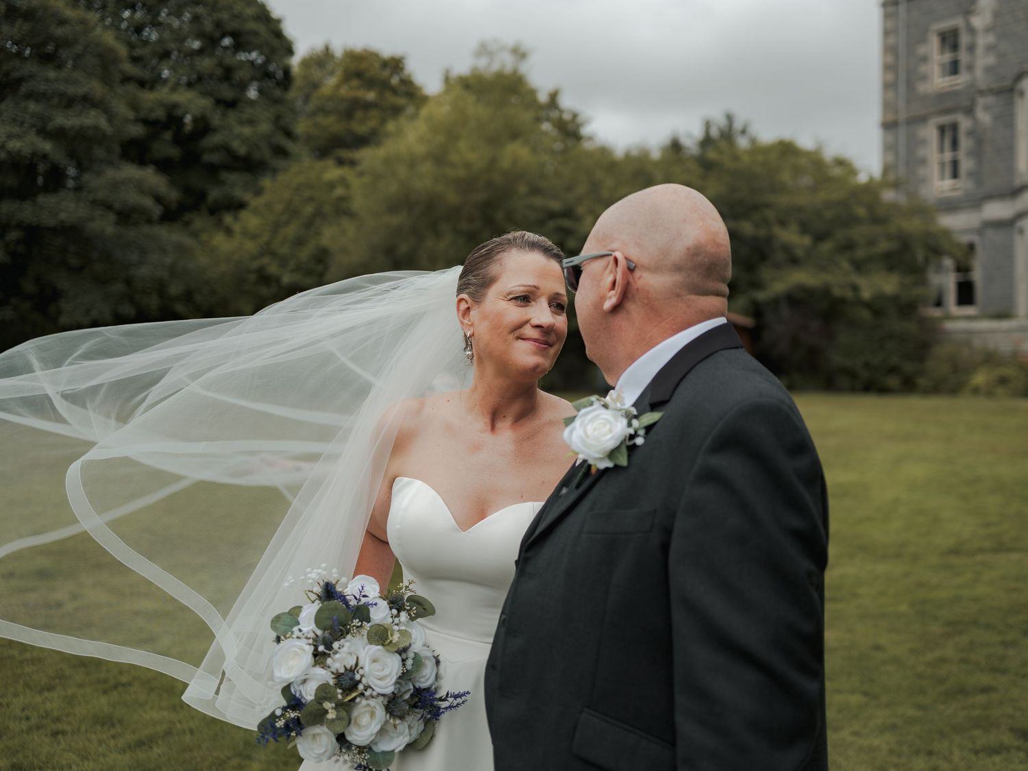 Bride and groom share a romantic moment outdoors, her veil flowing in the wind, holding a white bouquet near a historic estate.
