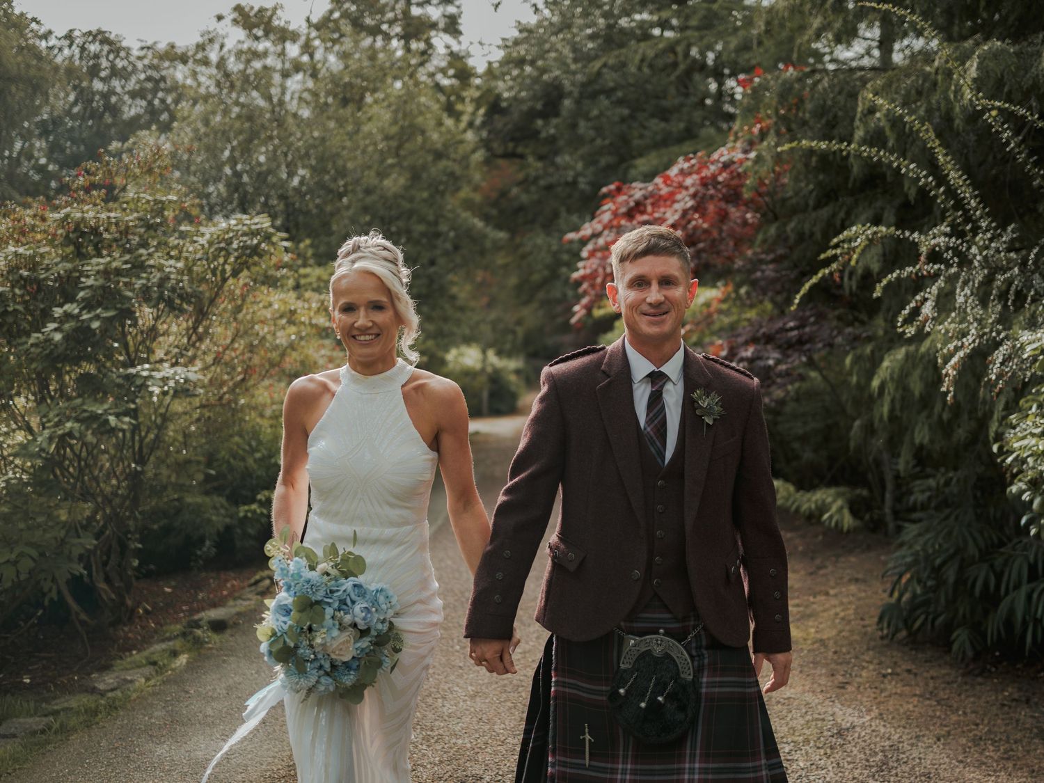Bride in white gown and groom in tartan kilt hold hands walking through a lush garden on their wedding day.