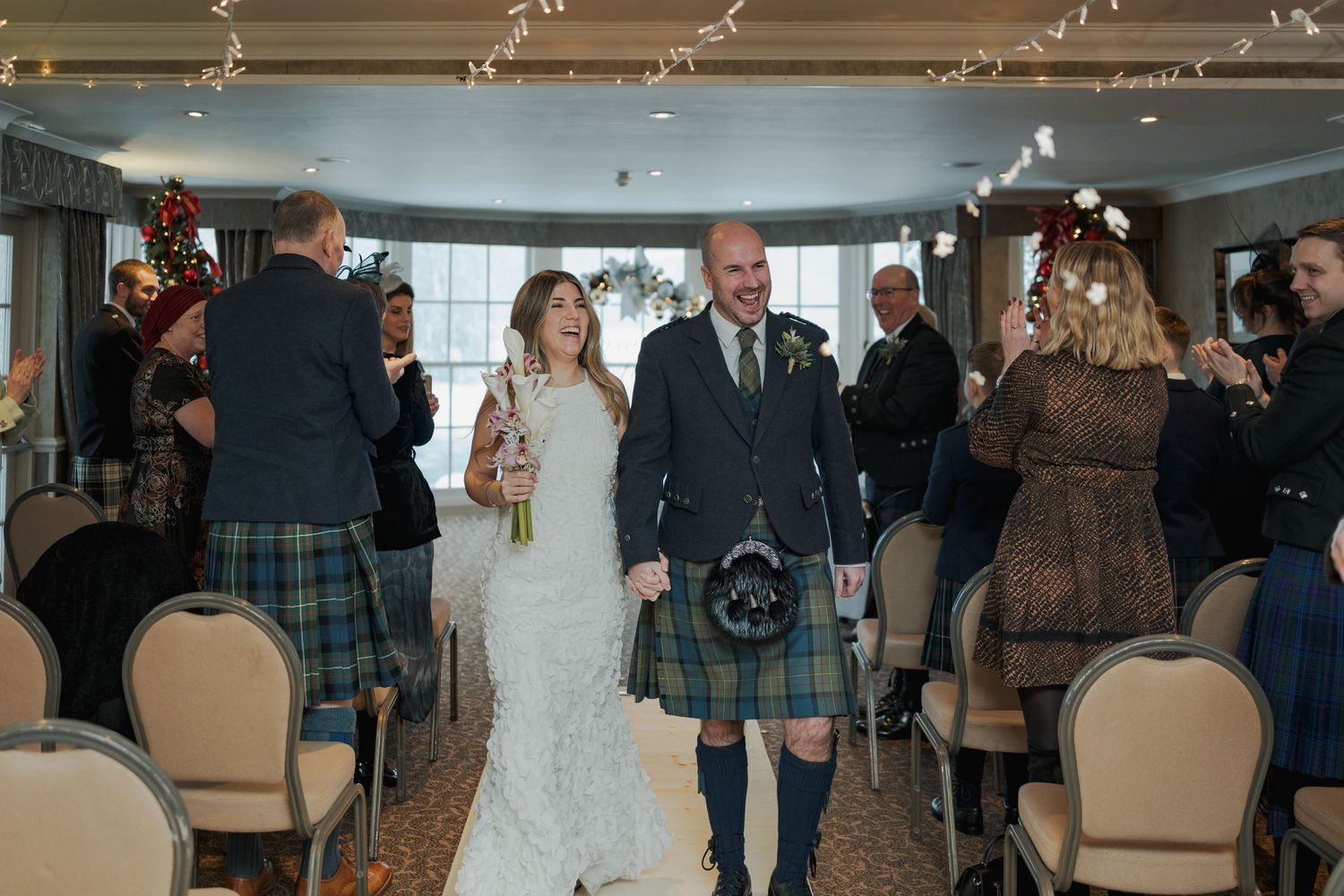 Newlyweds walk down aisle as guests celebrate, groom wearing traditional Scottish tartan kilt.