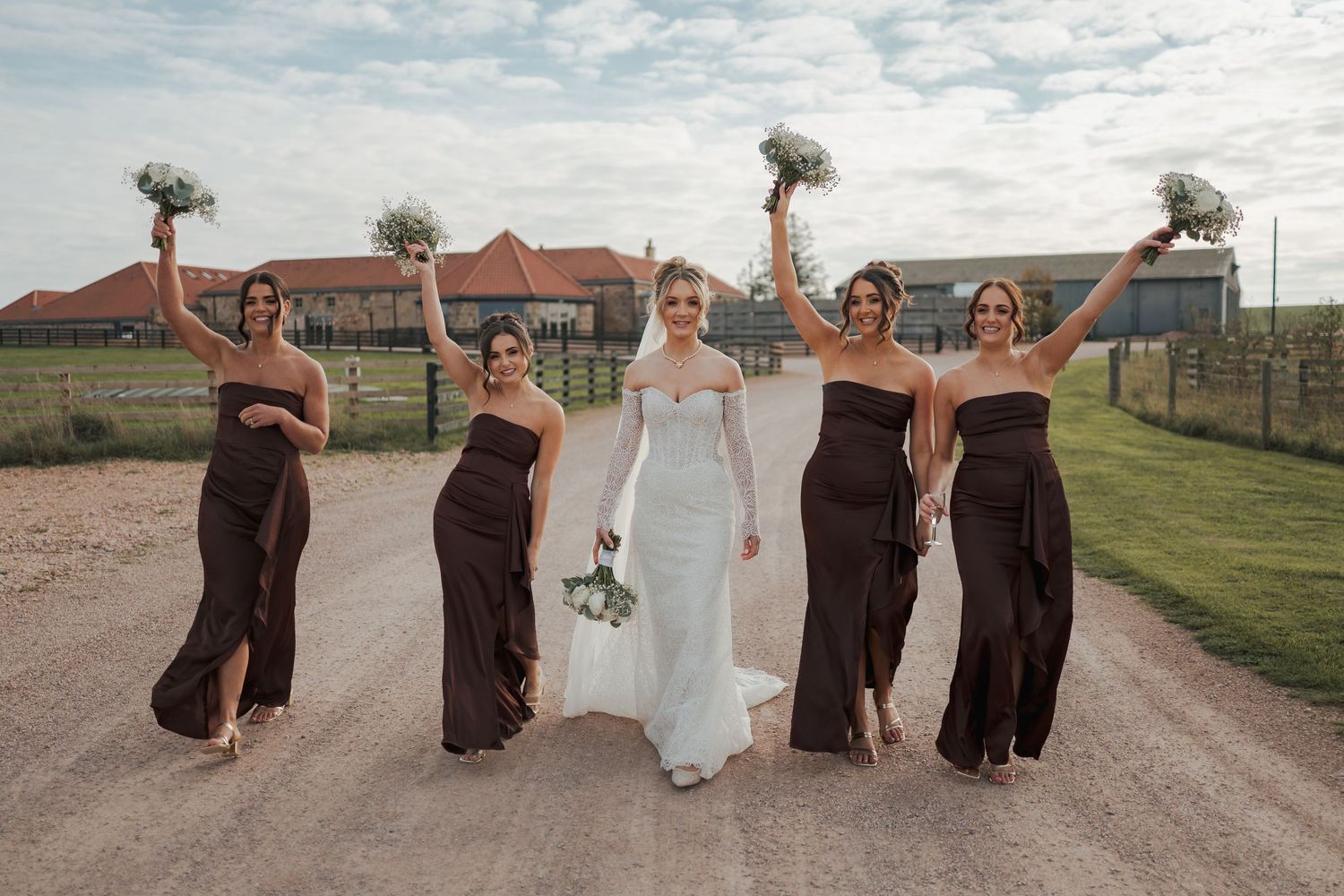 Wedding party in brown dresses and white gown walk down a road together at Falside Mill