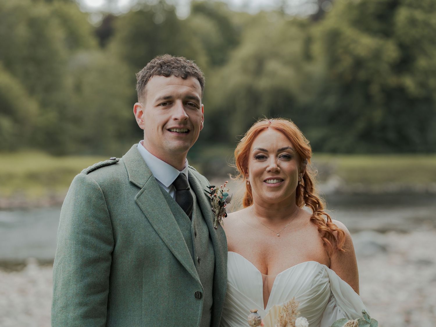 A couple shares laughter and smiles in intimate wedding portraits by a waterfront while wearing sage green attire at Banchory Lodge.