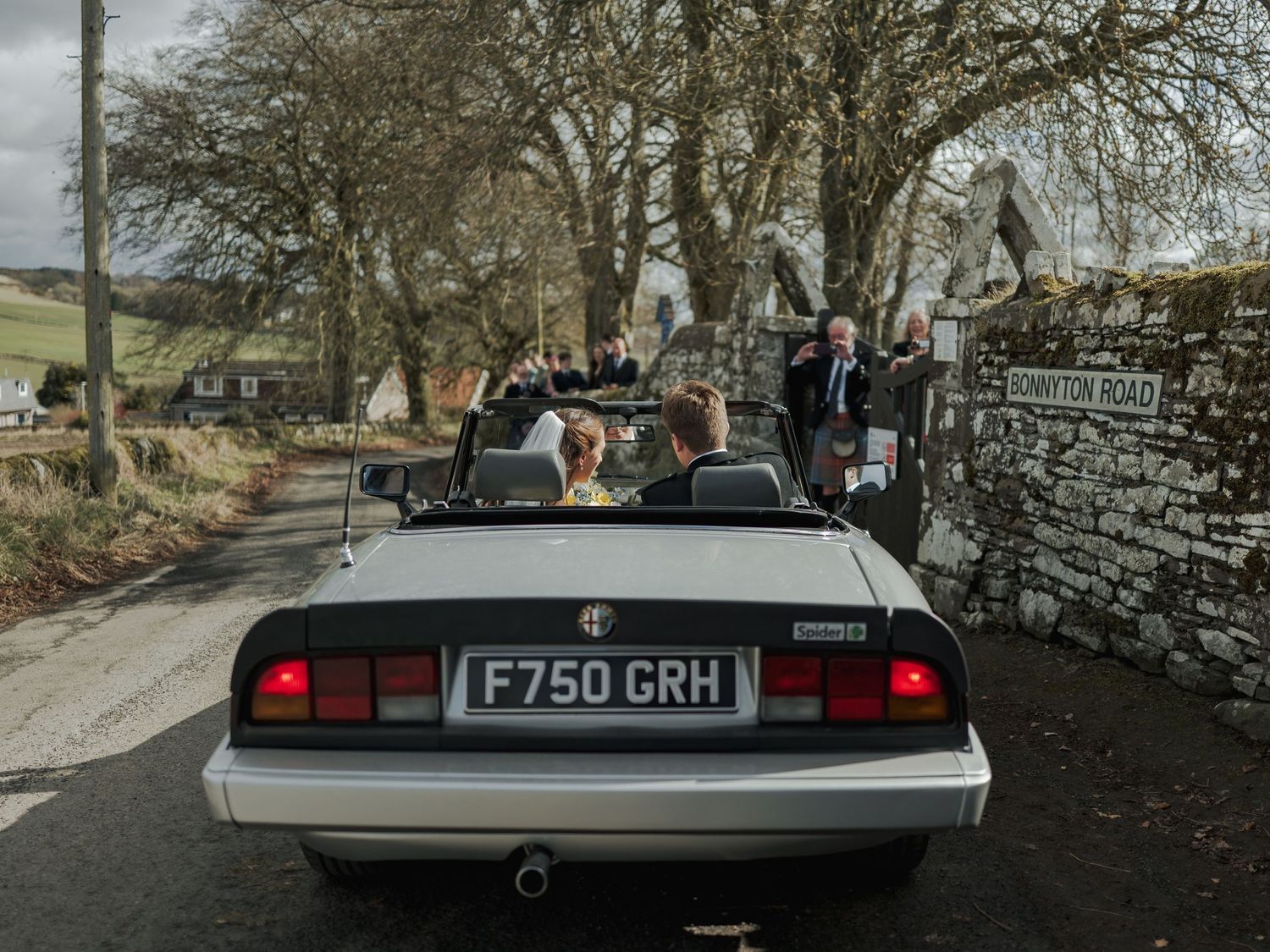 Wedding party riding in a vintage Alfa Romeo convertible on a rural road, license plate F750 GRH, near Tonton Road sign.
