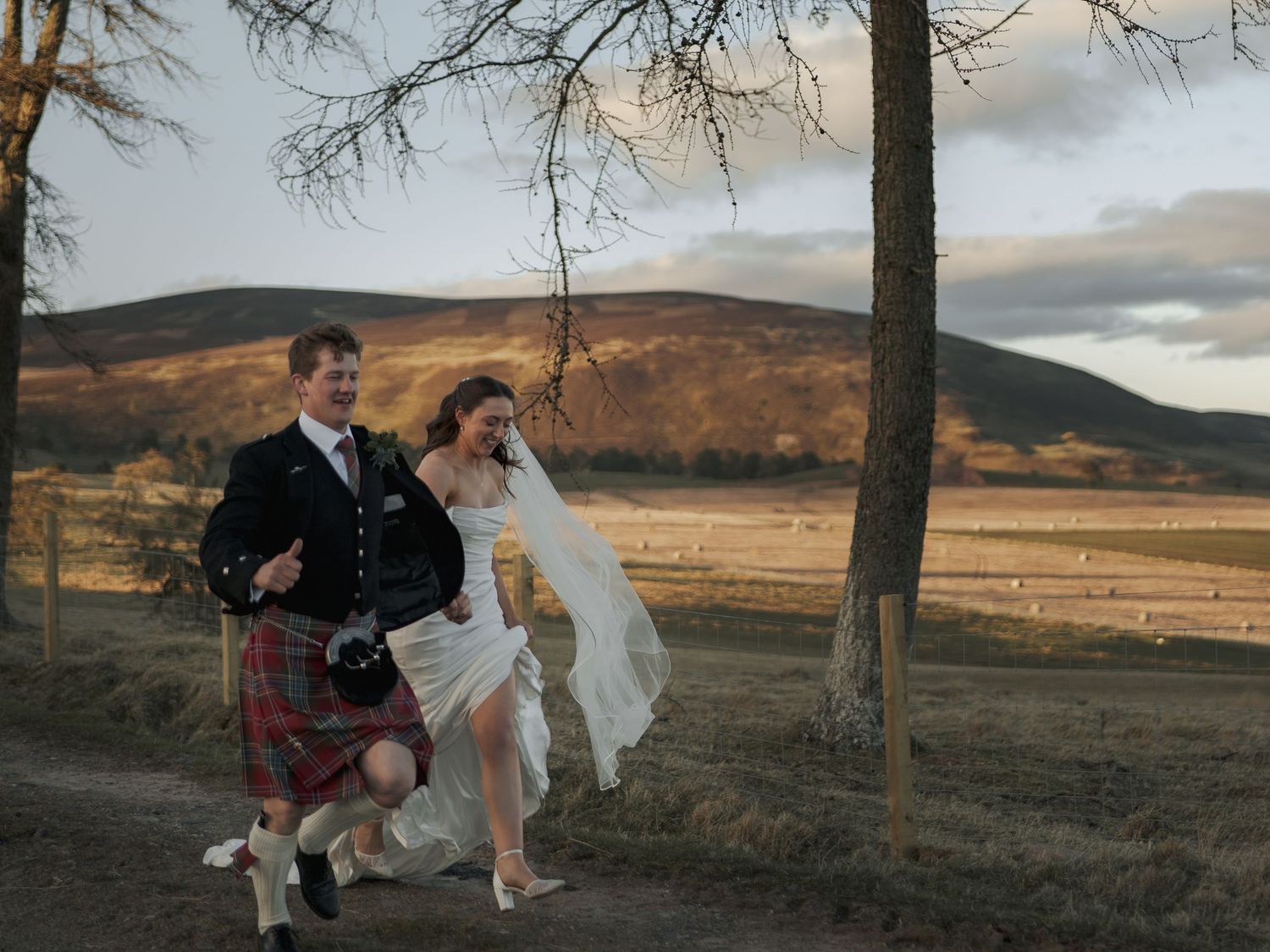 A joyful couple in Scottish wedding attire, groom in kilt, runs through a scenic Highland landscape at golden hour.