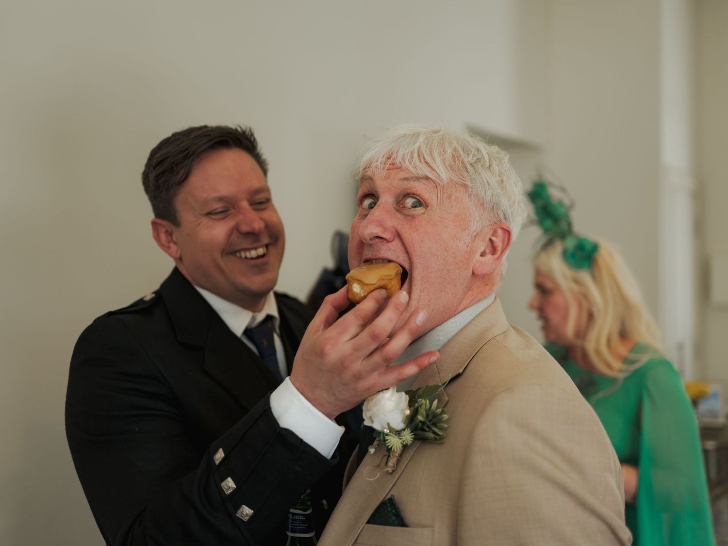 Groom laughing as best man feeds him a bite of food at a wedding reception celebration.