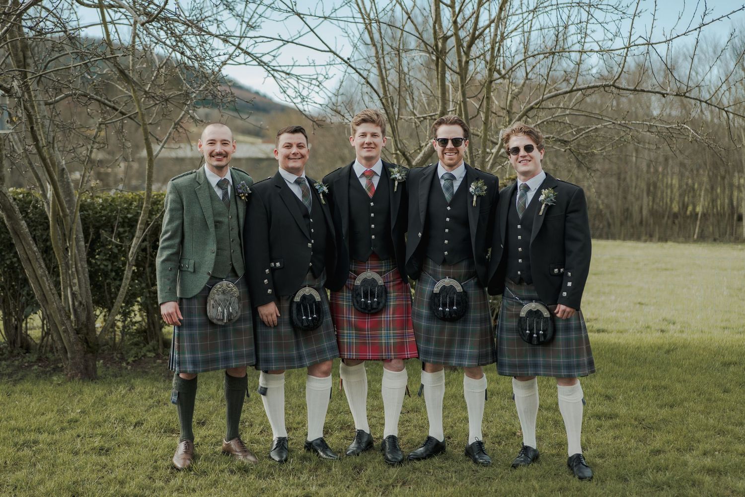Five men in traditional Scottish kilts and formal jackets pose outdoors at a wedding celebration.