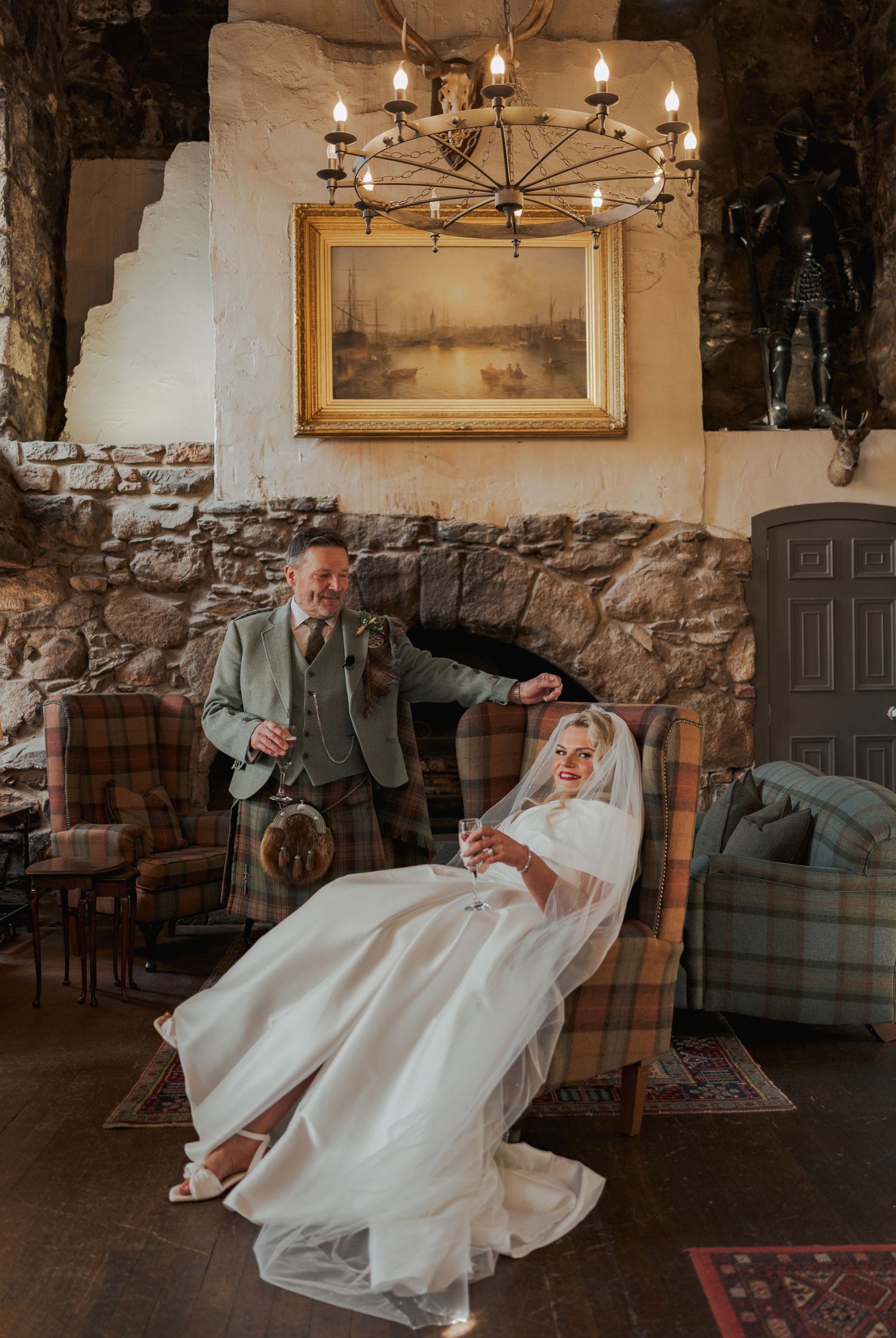 Bride in white gown seated by stone fireplace with man in Scottish kilt in a rustic castle interior.