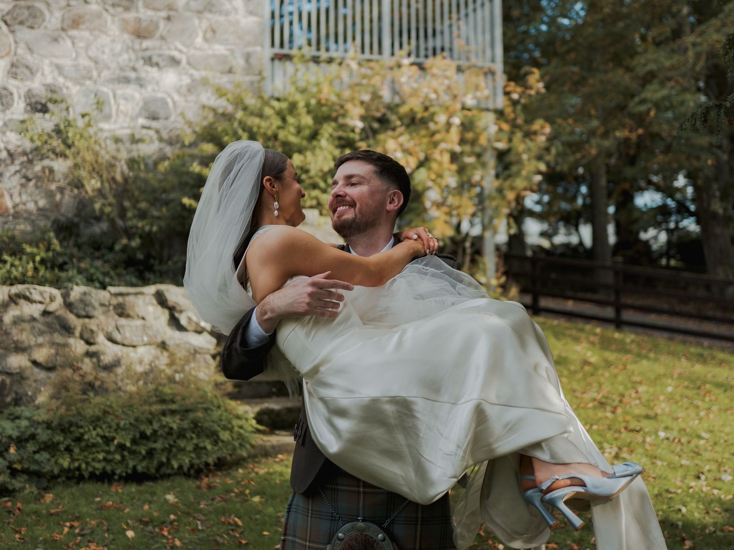 Groom in kilt carrying bride in white wedding dress and veil outdoors near stone wall with autumn trees.