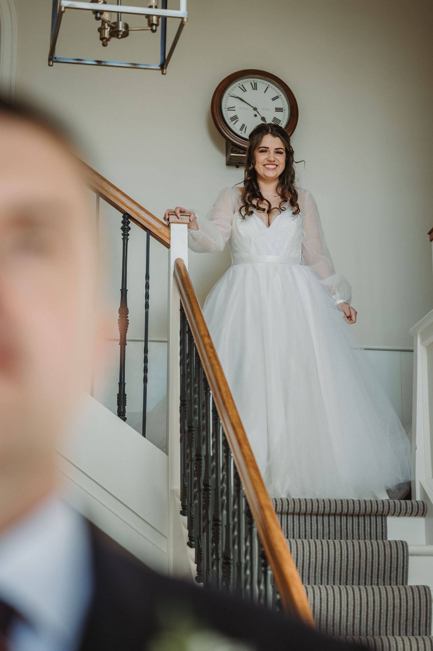 A bride in a long white wedding dress stands joyfully on carpeted stairs under a classic wall clock at Kinclune House.