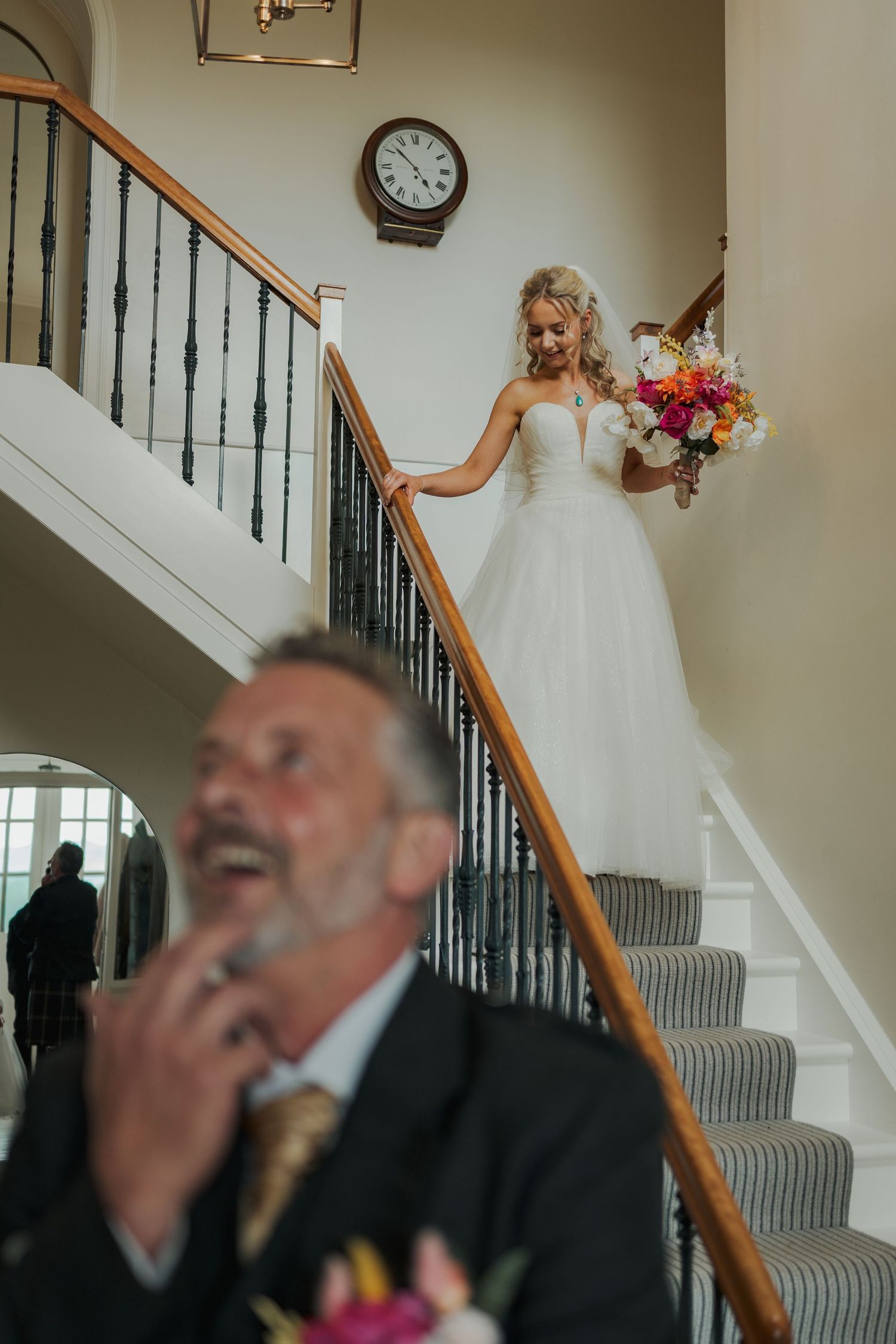 Bride in white gown descends staircase holding colorful bouquet as emotional father waits in foreground.