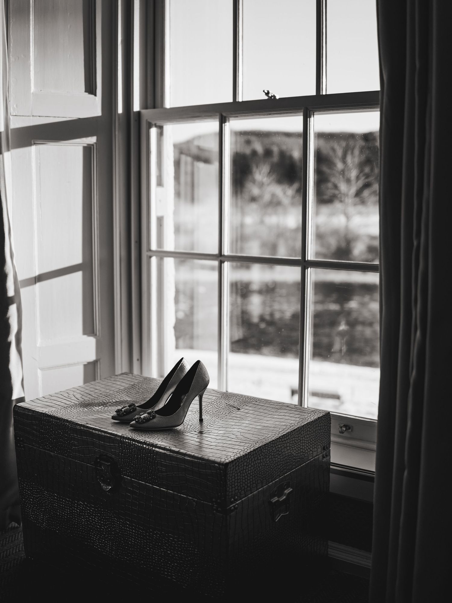 Black and white photo of elegant high heel shoes on a vintage trunk by a window with trees outside.