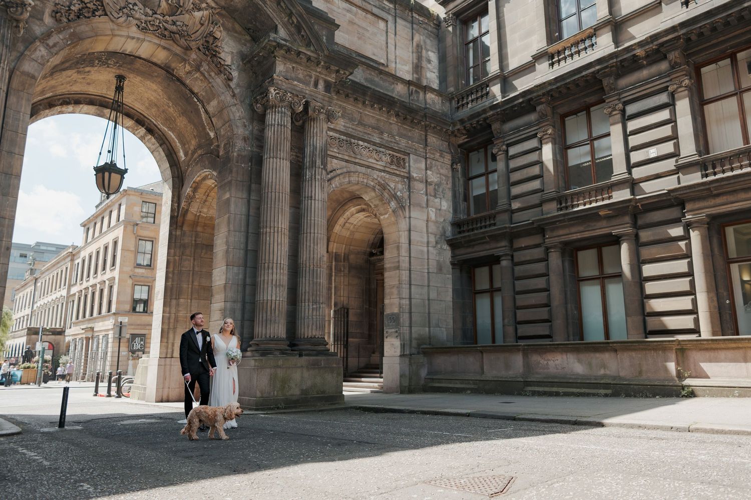 Historic stone buildings and archways line a cobblestone street in Glasgow's merchant quarter on a cloudy day.