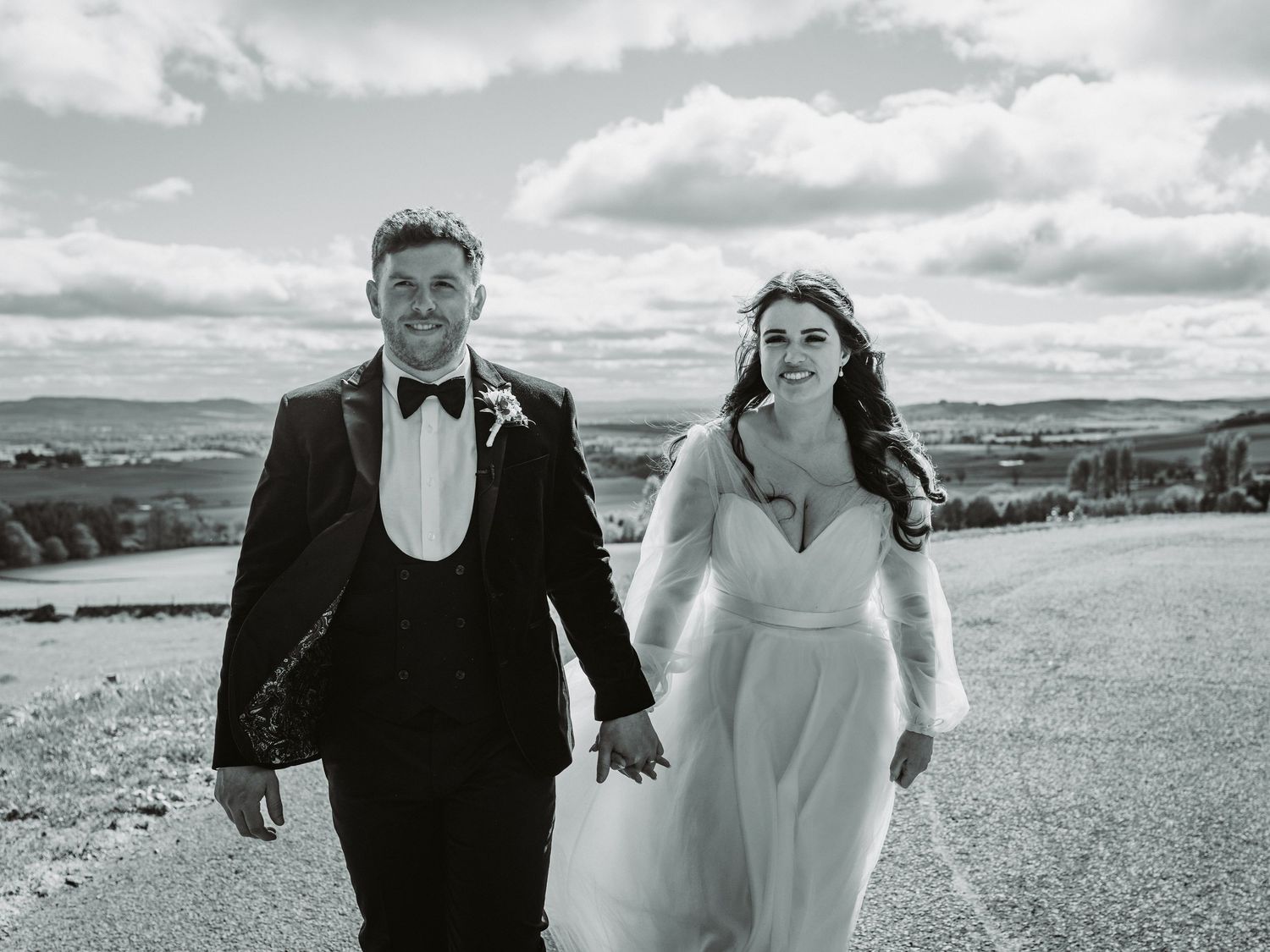 A couple in formal wedding attire walk hand in hand on a hilltop path with dramatic cloudy sky and countryside views across Kirriemuir.