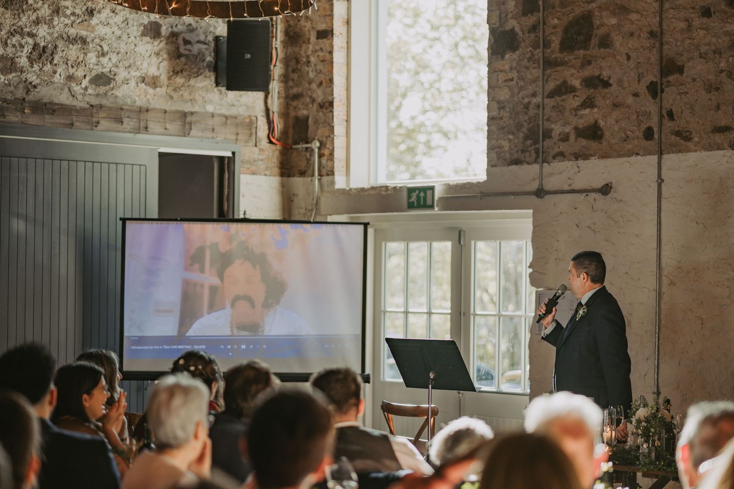 Father of the bride presents to an audience in the rustic Kinclune Steading with exposed brick walls and a large screen display.