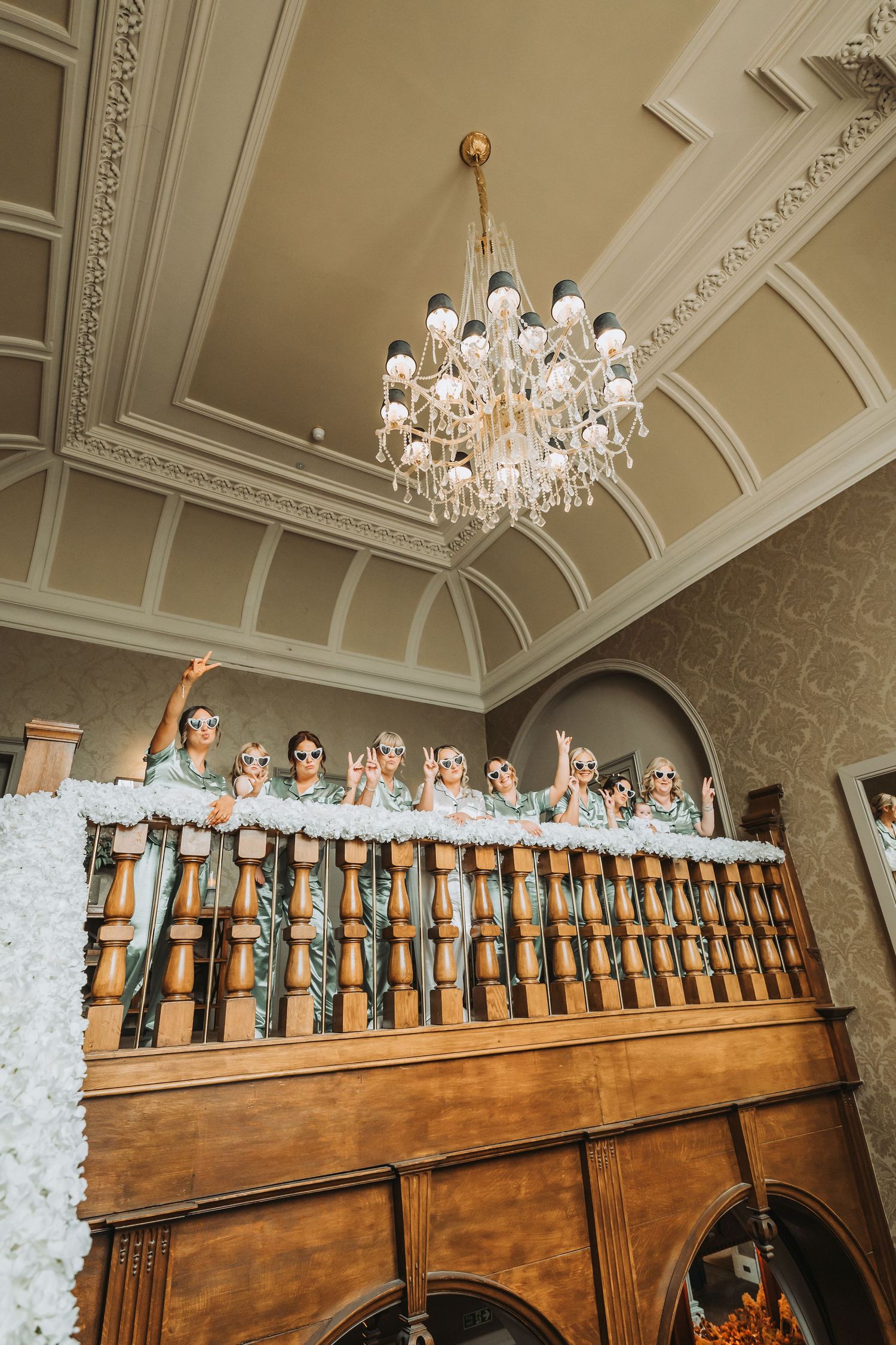 Wedding party poses along elegant wooden balcony under crystal chandelier in historic venue Cornhill Castle.