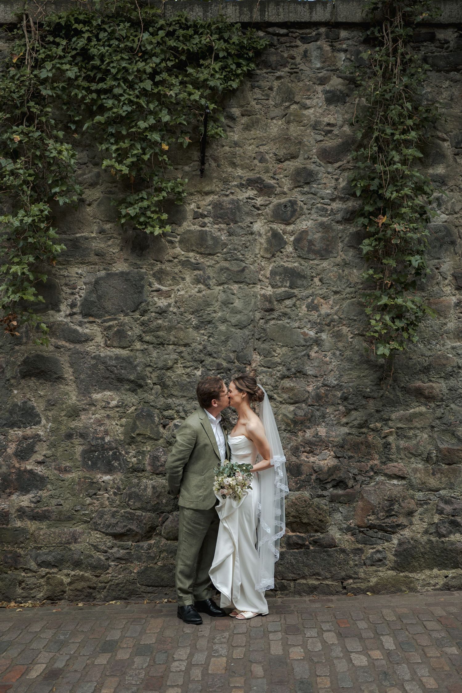 Bride and groom kissing against a rustic stone wall with ivy, bride holding a bouquet in a white gown and veil.