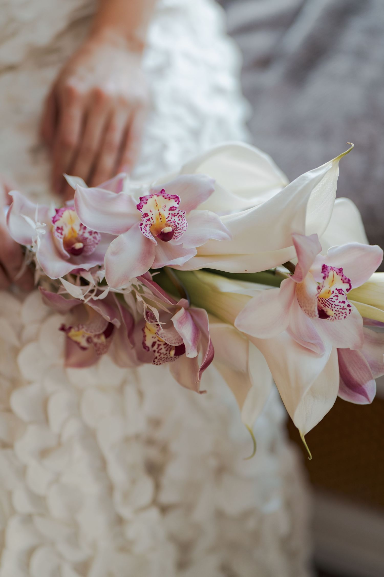 Bride holding elegant wedding bouquet of white calla lilies and pink cymbidium orchids against a textured white lace gown.