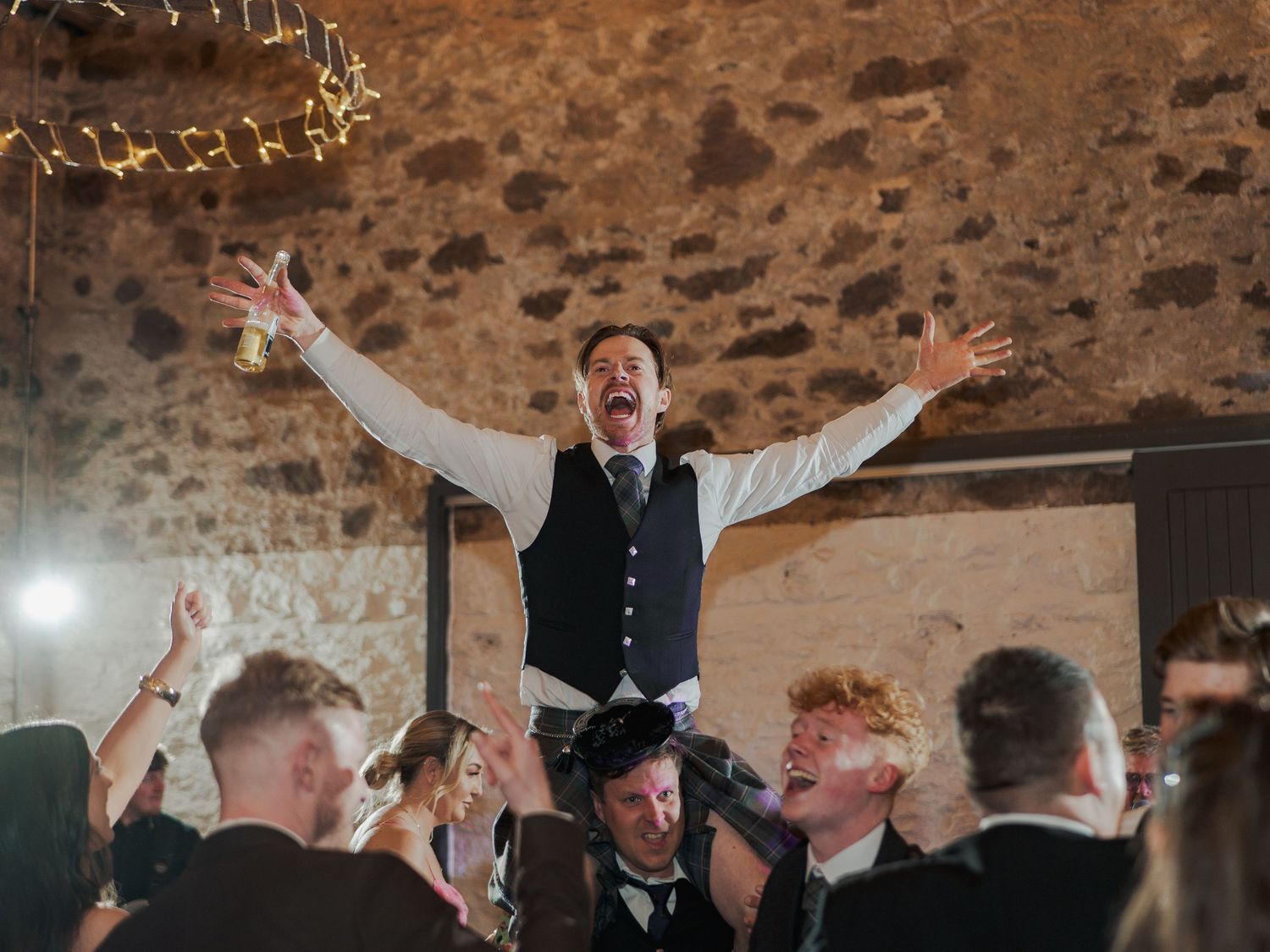 Groom celebrating with arms raised at wedding reception in rustic stone venue with cheering guests.