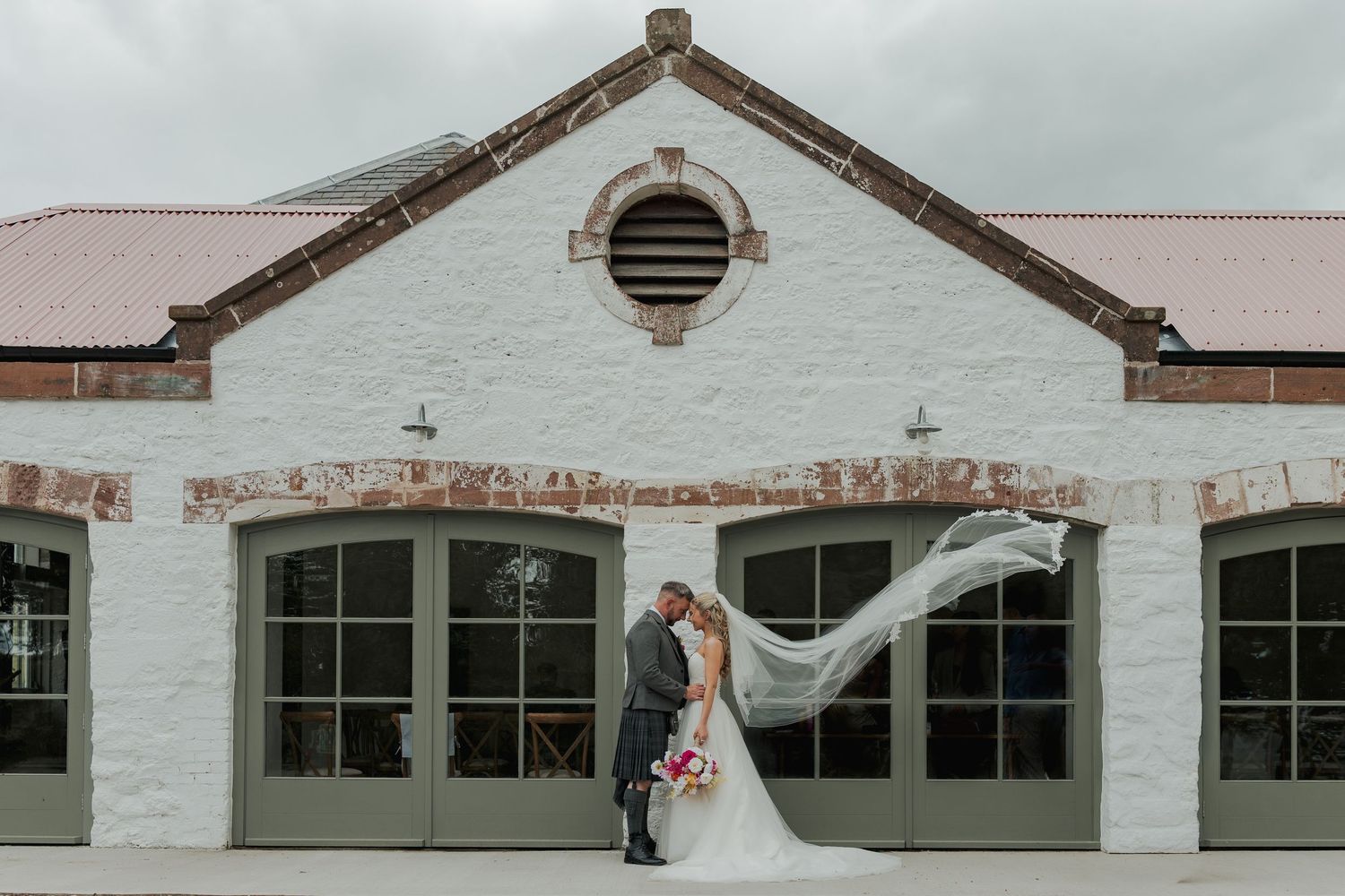 Bride and groom kissing outside a white historic building with veil flowing in the wind on their wedding day.