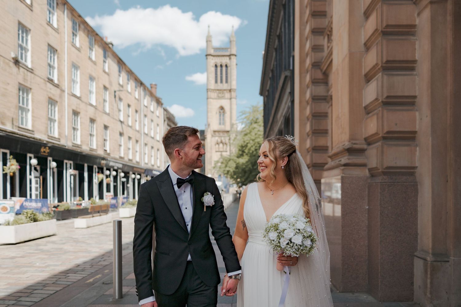 A couple in wedding attire walks through a grand stone colonnade with ornate architecture.