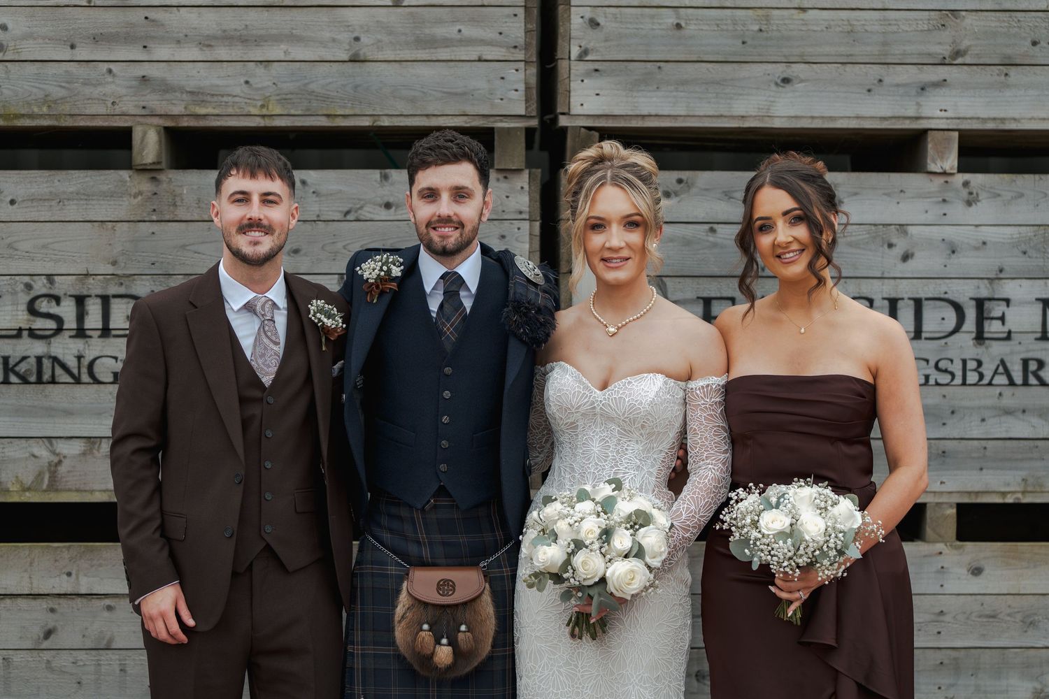 Wedding party members pose together in front of rustic wooden wall wearing formal attire and holding bouquets at Falside Mill.