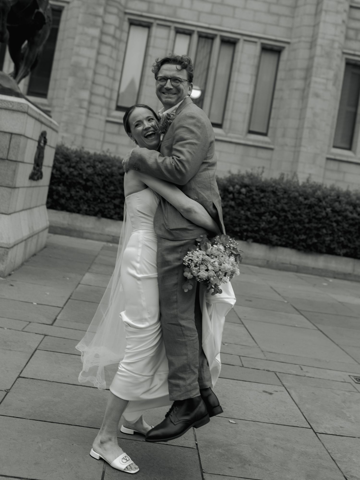 Joyful newlyweds embrace and laugh outside a stone building, bride in white gown holding bouquet, black and white photo.