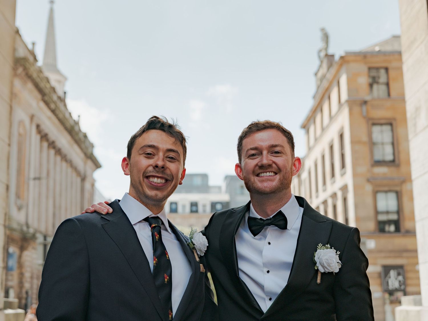 A wedding party in formal attire stands together smiling in a row against a backdrop of elegant stone buildings.